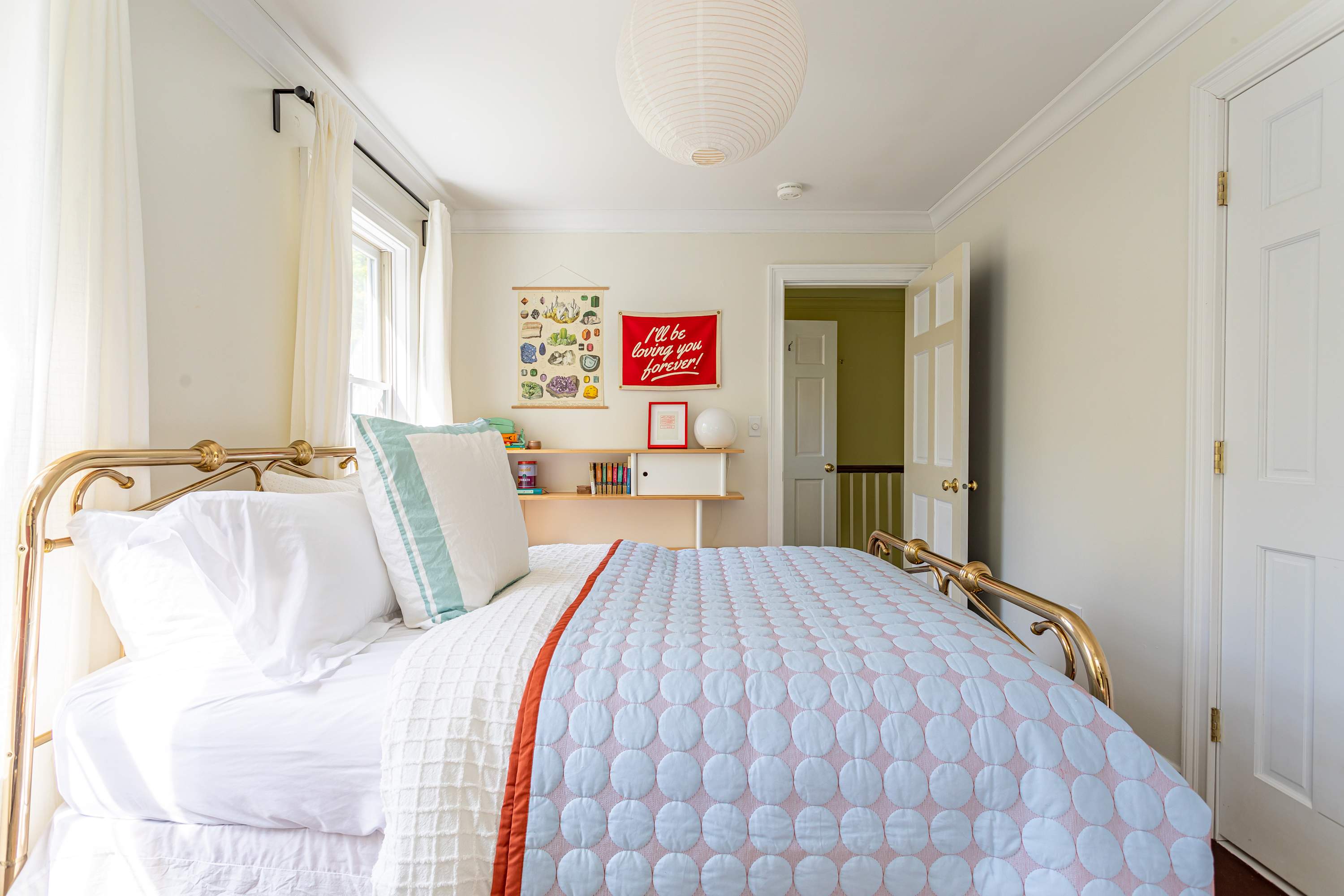 A cozy bedroom with a brass bed frame and a blue and white quilted comforter, facing a doorway that leads to a hallway. A white paper lantern hangs from the ceiling, and a built-in shelf unit is visible on the wall.