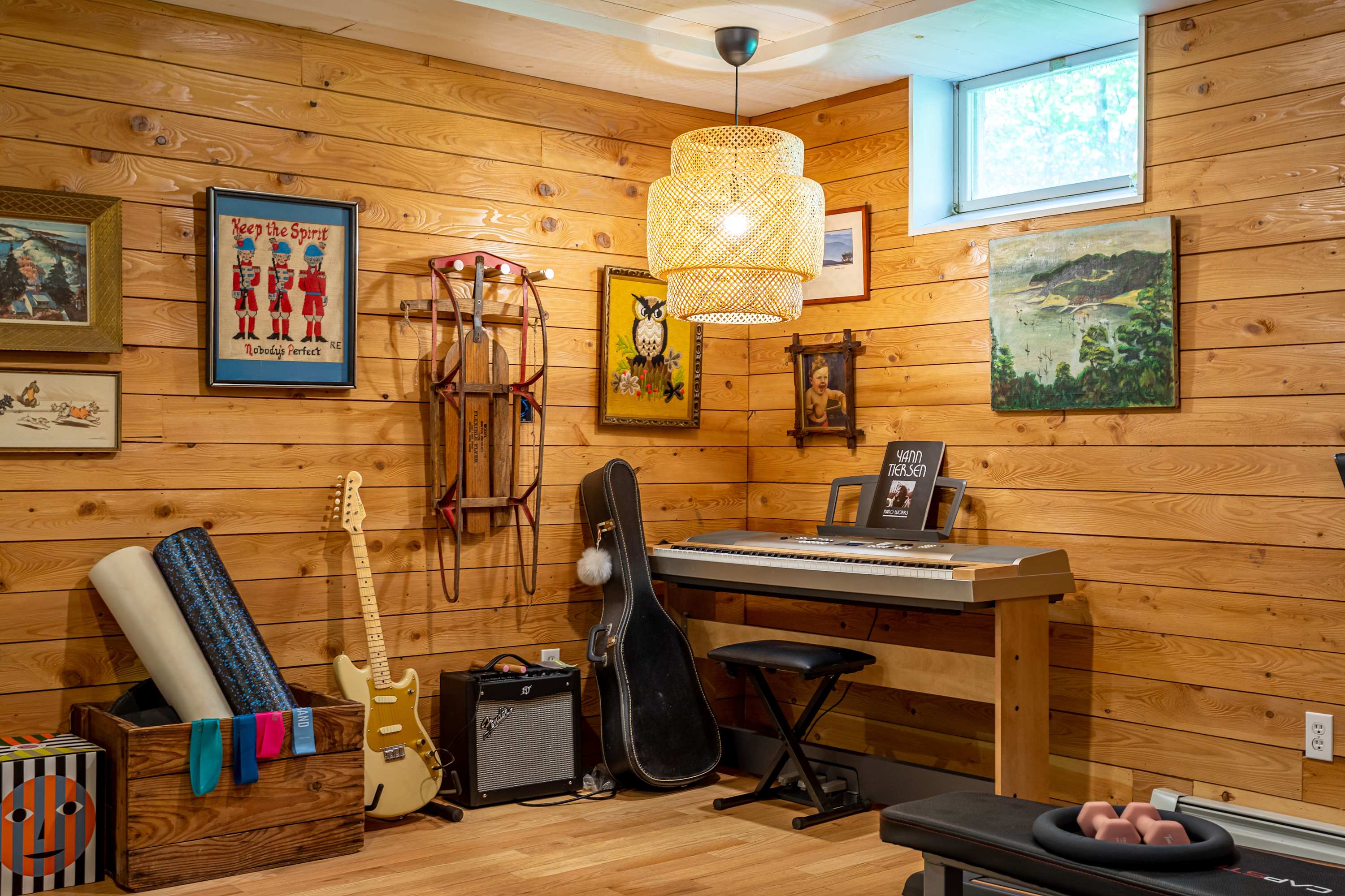 Multi-functional recreation room with rustic wood-paneled walls and light hardwood floors, showcasing an electronic piano, a guitar with an amplifier, and various exercise equipment. Eclectic artwork and a decorative sled adorn the walls, illuminated by a woven pendant light.