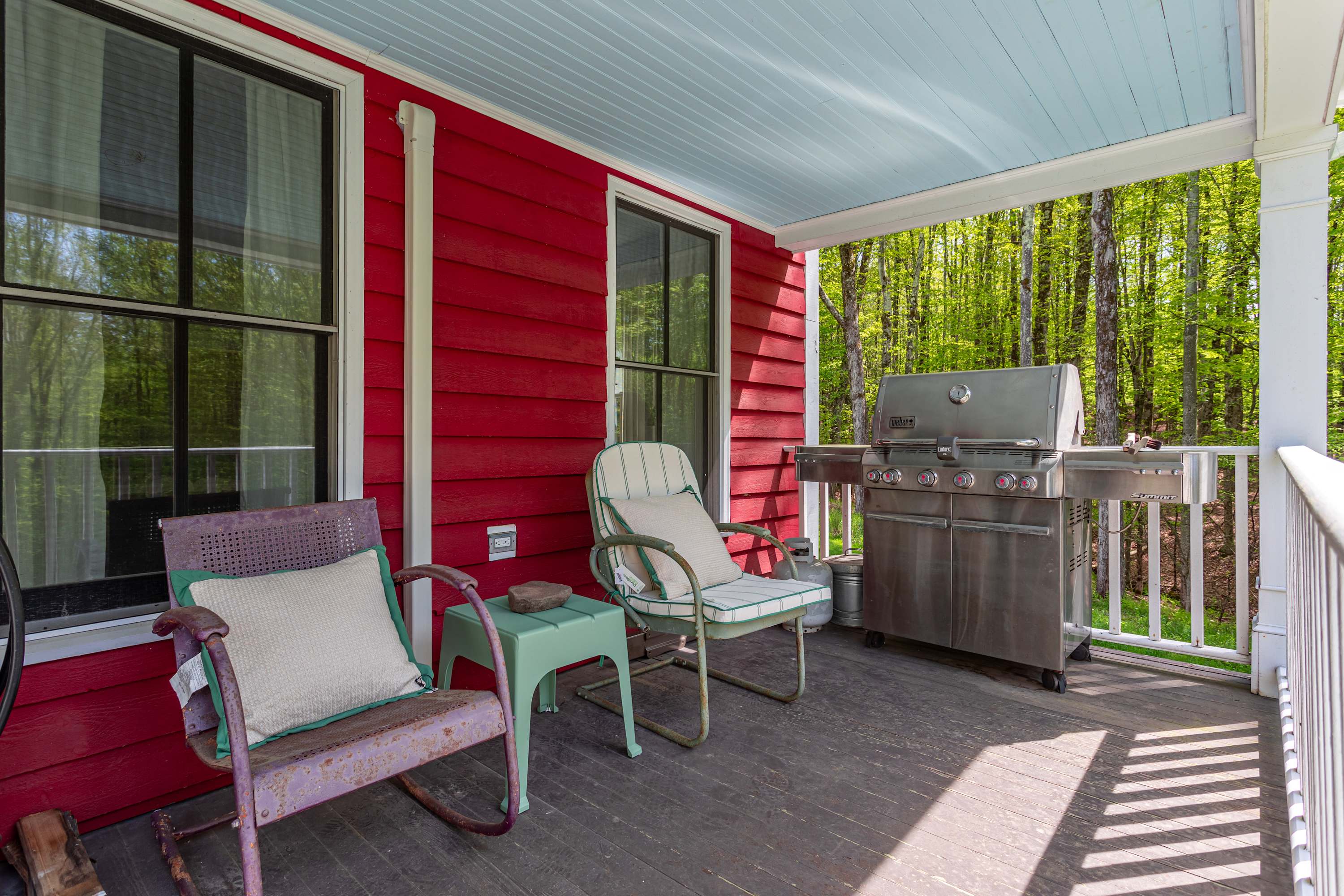 Relaxing covered porch with a light blue ceiling and dark wood flooring, furnished with two vintage-style chairs and a small side table. A large stainless steel gas grill is positioned near the railing, overlooking a lush green forest.