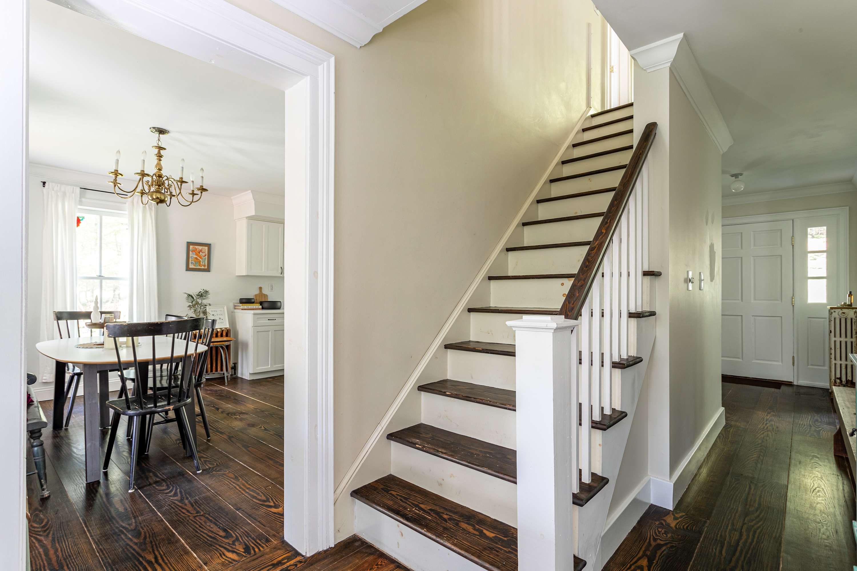 A welcoming foyer with dark hardwood floors and a white staircase featuring dark wood treads and a white banister. An archway leads to a dining area with a round table and black chairs, illuminated by a chandelier.
