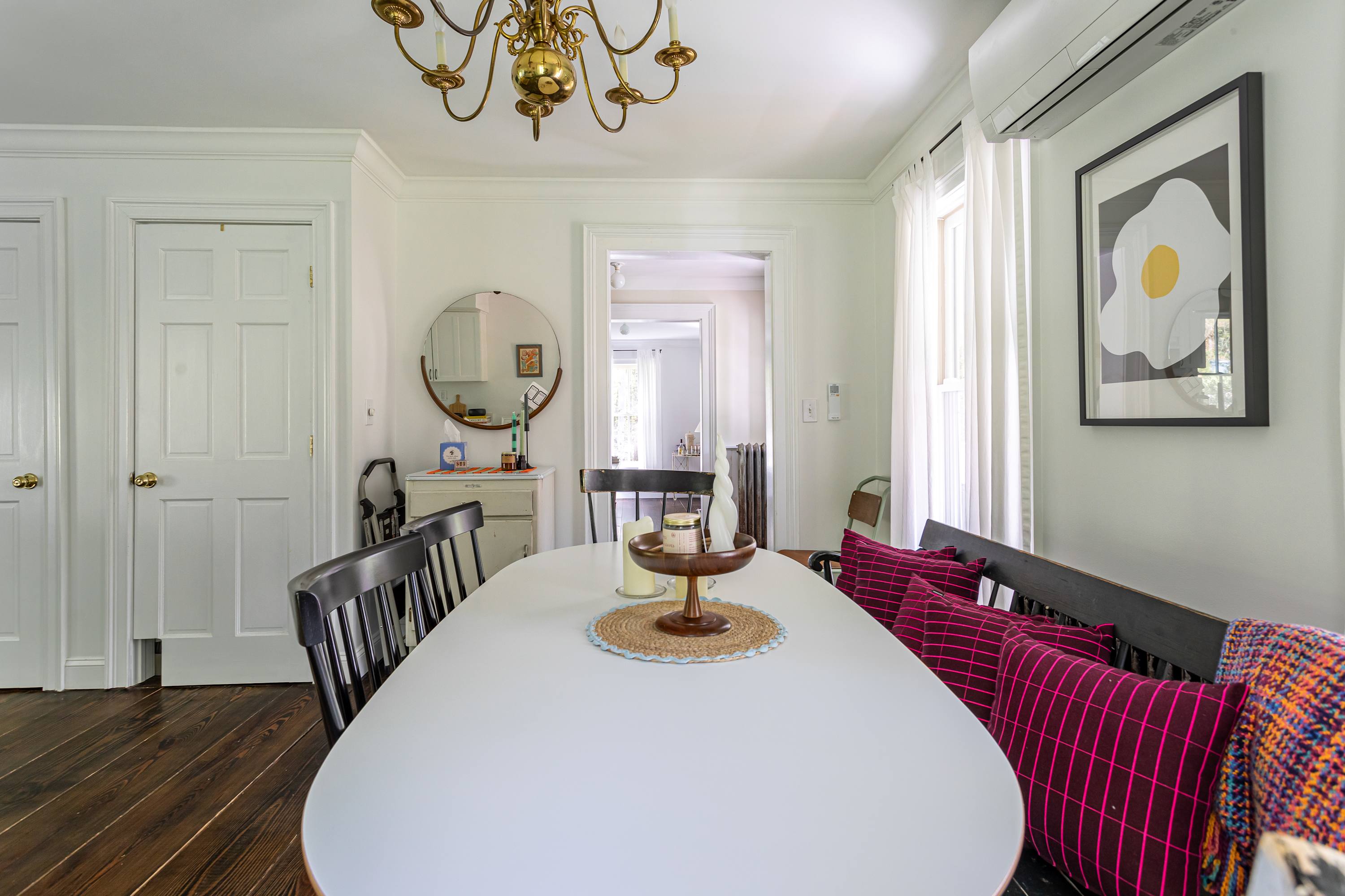 Bright dining area with a long white oval dining table, black chairs, and a black bench with magenta pillows. Dark hardwood floors and white walls are complemented by a brass chandelier and a round mirror.