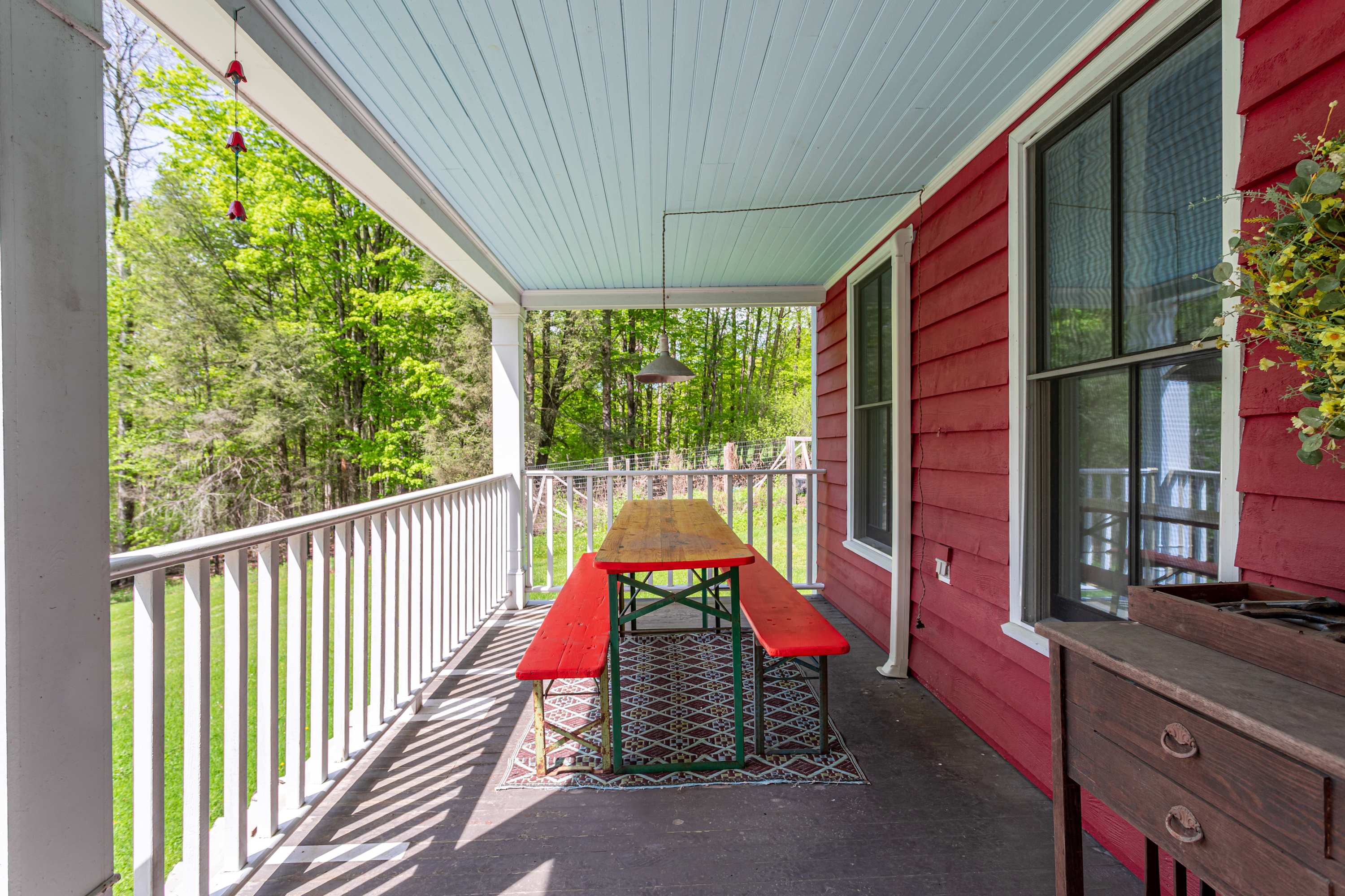 Inviting covered front porch with a light blue ceiling and dark wood flooring, featuring a long wooden picnic table with red benches. White railings line the porch, offering a view of the surrounding green trees.