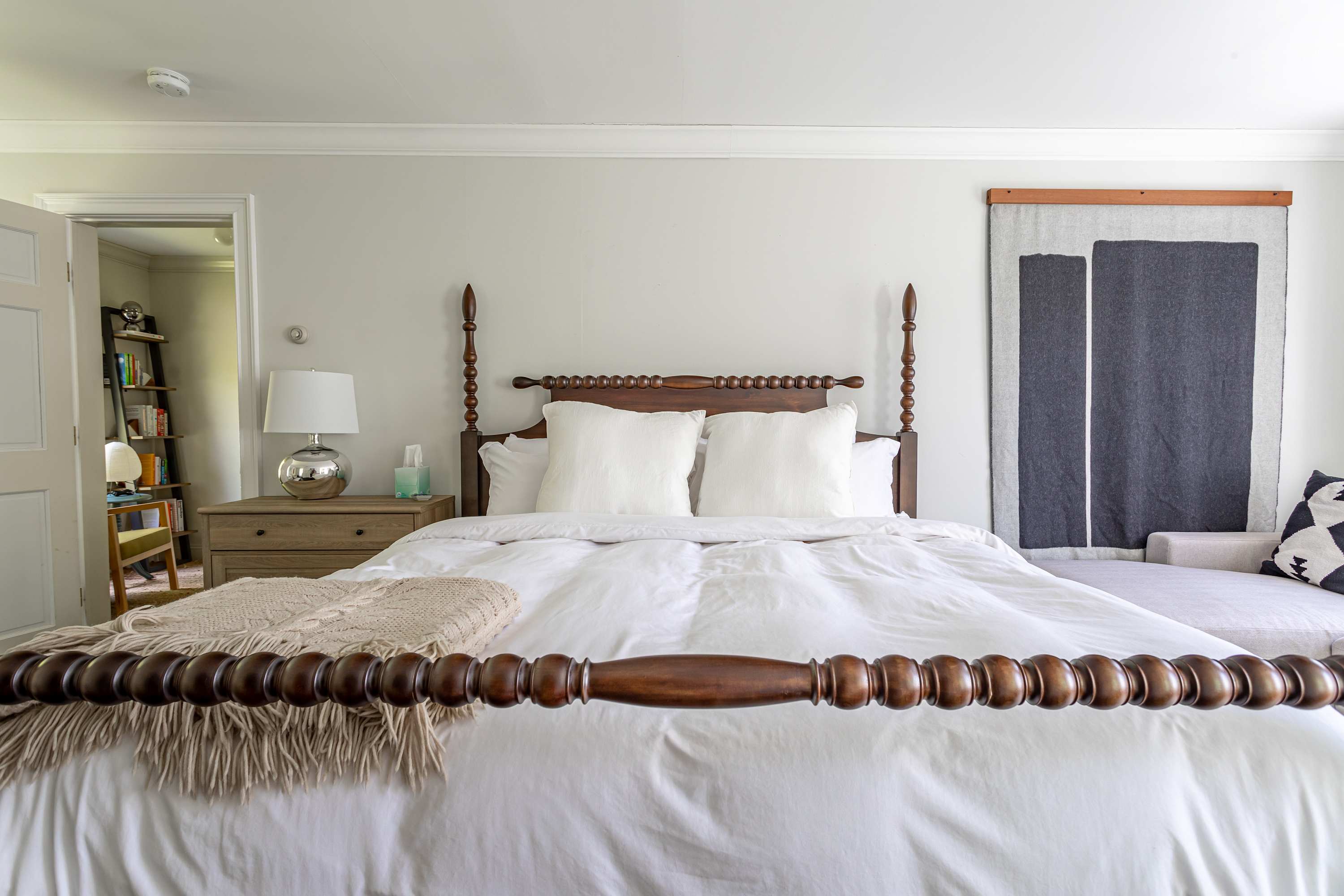 A wide shot of the bedroom with a dark wood four-poster bed, white bedding, and a large abstract wall hanging. A doorway leads to an adjacent room, and a sitting area with a sofa is visible near the window.