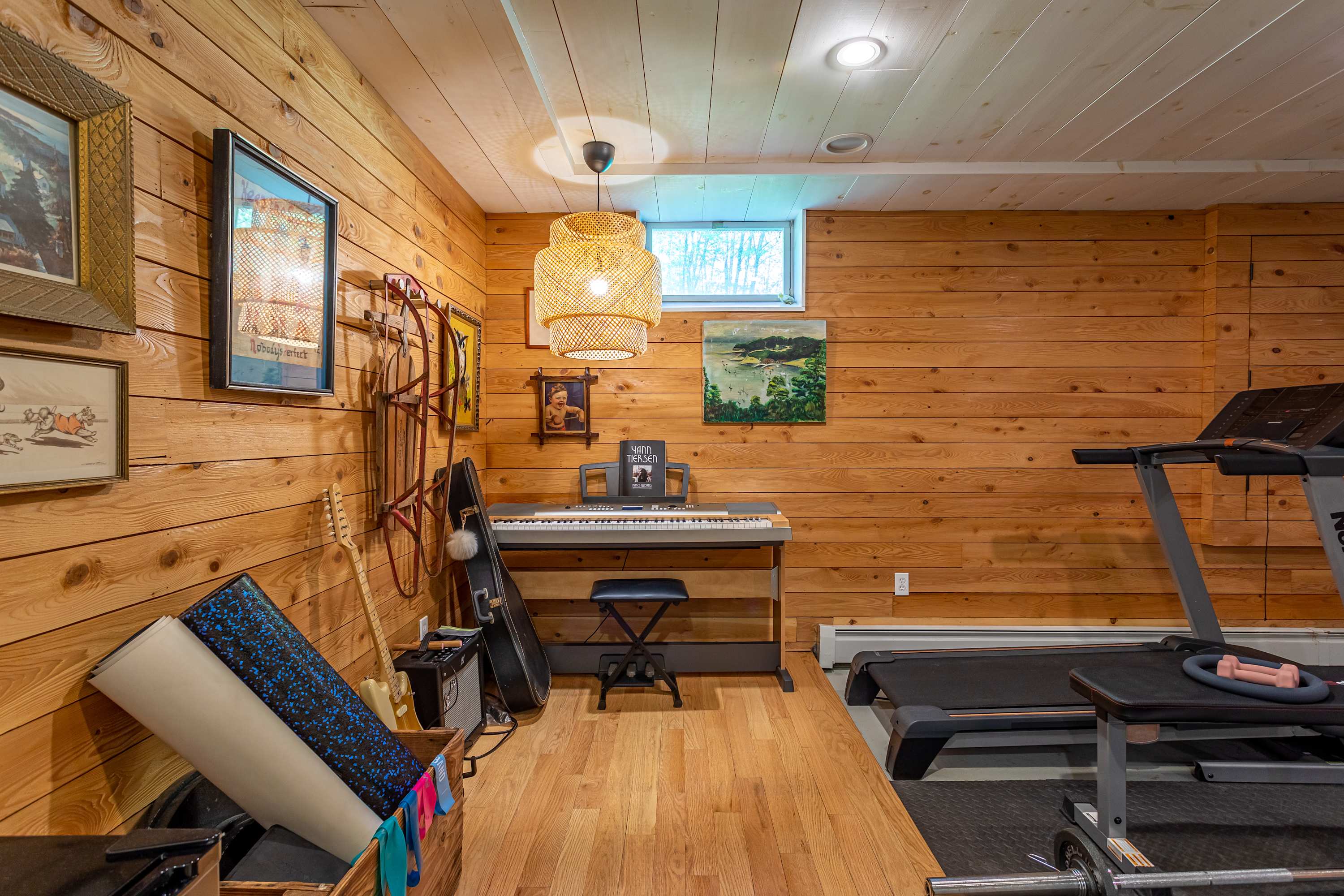 Cozy recreation room with warm wood-paneled walls and light hardwood floors, featuring an electronic piano with a stool and a treadmill. A small window is visible high on the wall, and a woven pendant light hangs from the ceiling.