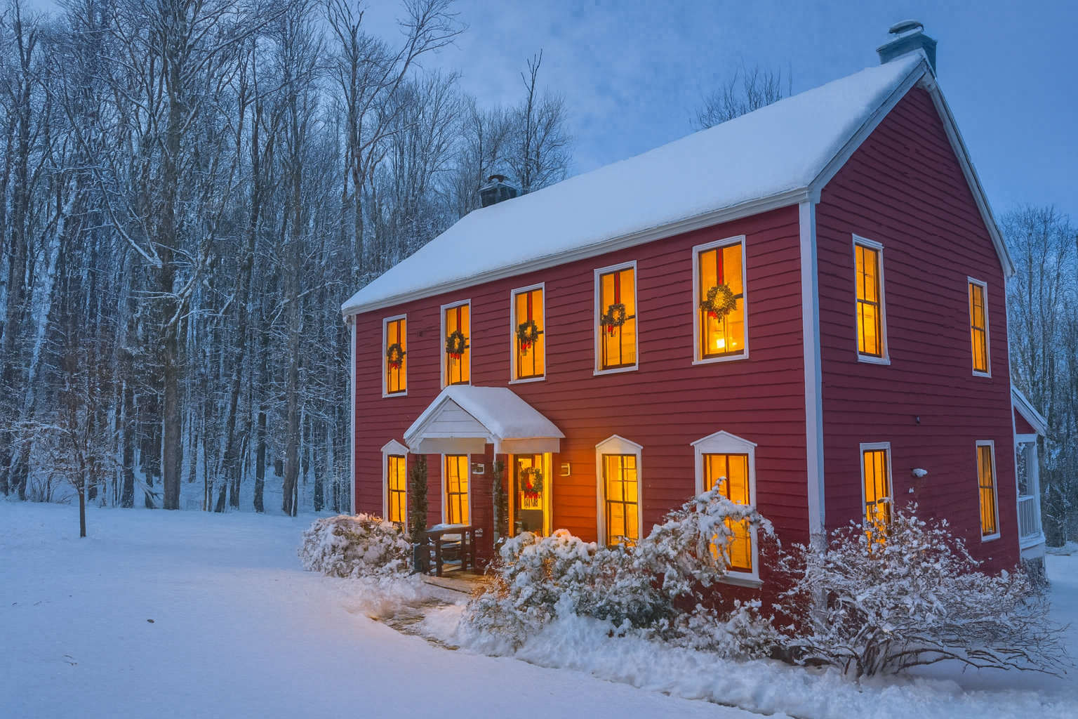 Picturesque red farmhouse blanketed in snow during winter, with warm lights glowing from inside each window adorned with festive wreaths. Bare trees stand in the background under a twilight sky, creating a cozy, holiday atmosphere.