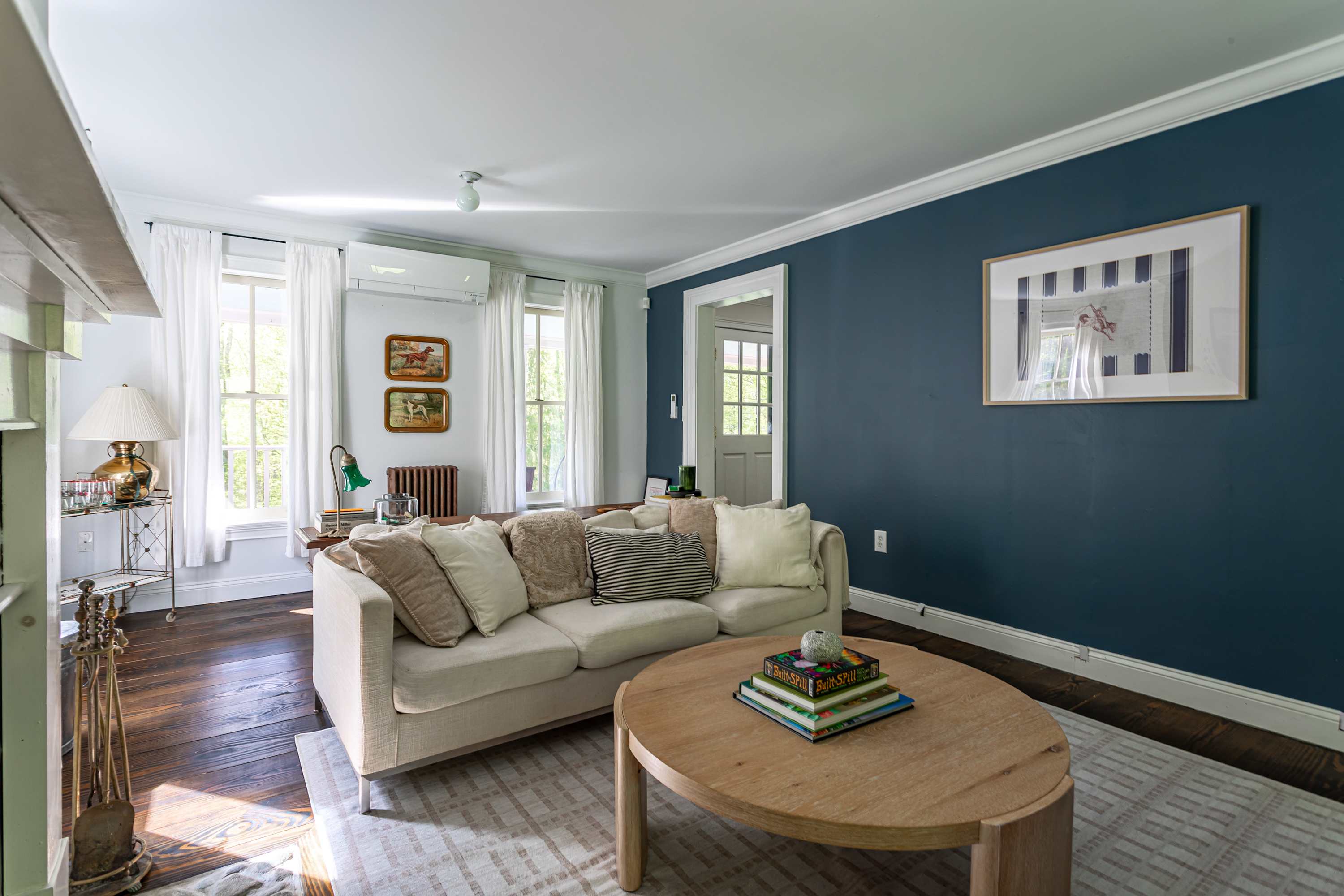 Bright living room with a cream sofa, a round wooden coffee table, and a striking dark blue accent wall. White sheer curtains adorn the windows, and dark hardwood floors run throughout.