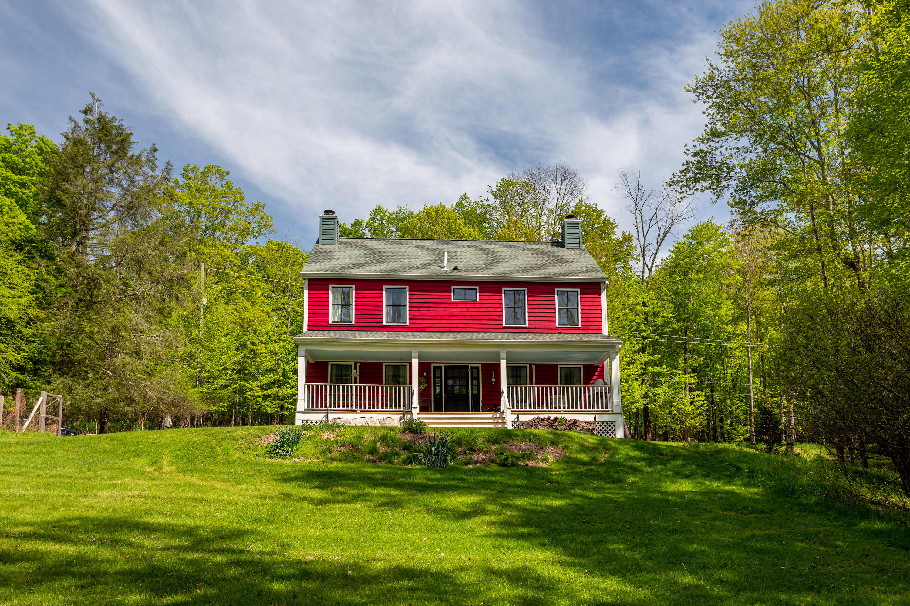 Charming red farmhouse with white trim and a covered front porch, situated on a lush green lawn surrounded by mature trees under a partly cloudy sky. The house features multiple windows and a classic, inviting facade.