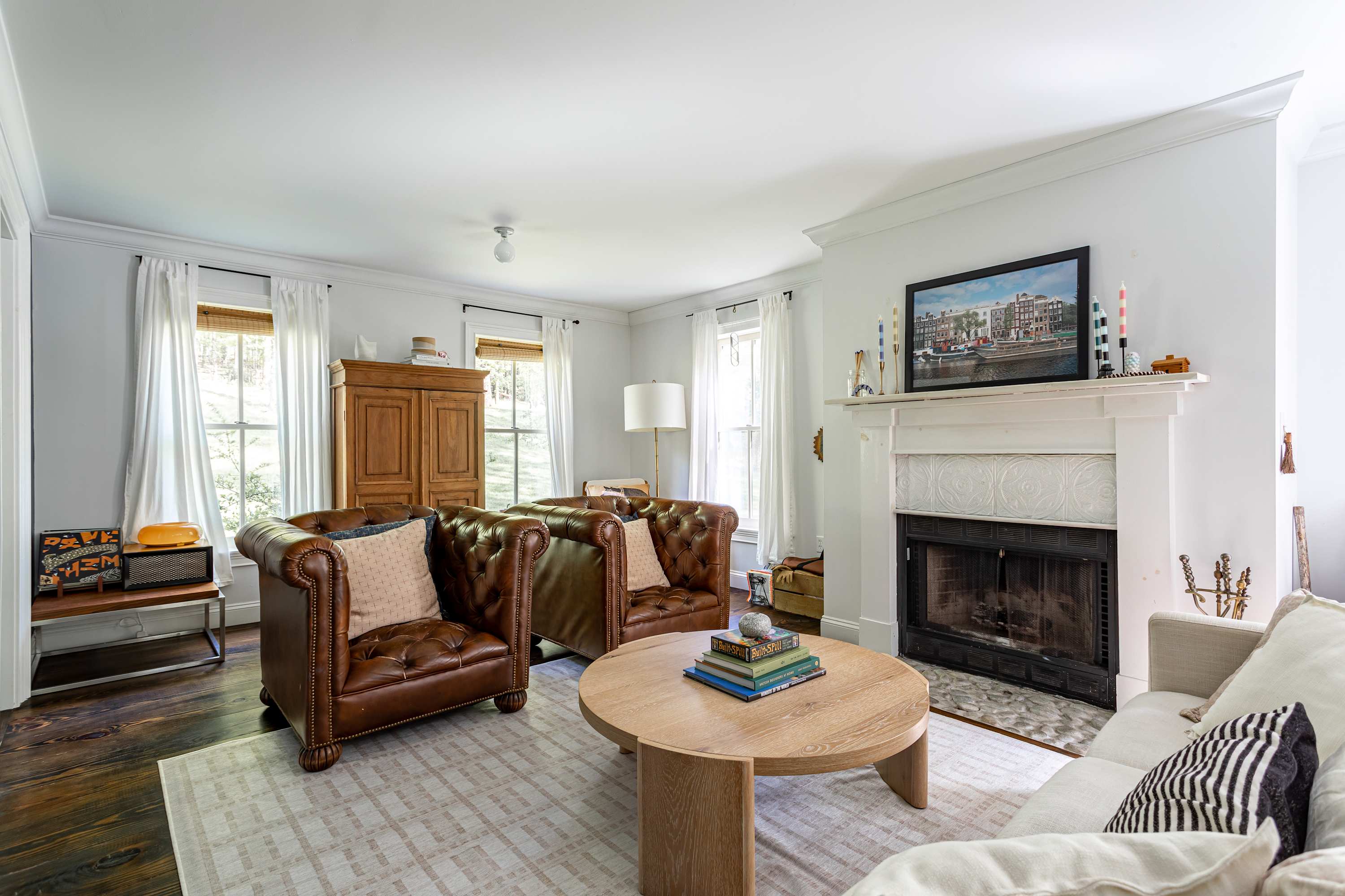 Inviting living room with two brown leather tufted armchairs, a white sofa, and a round wooden coffee table on a light patterned rug. A white fireplace with a mantel and a large window with bamboo blinds are visible.