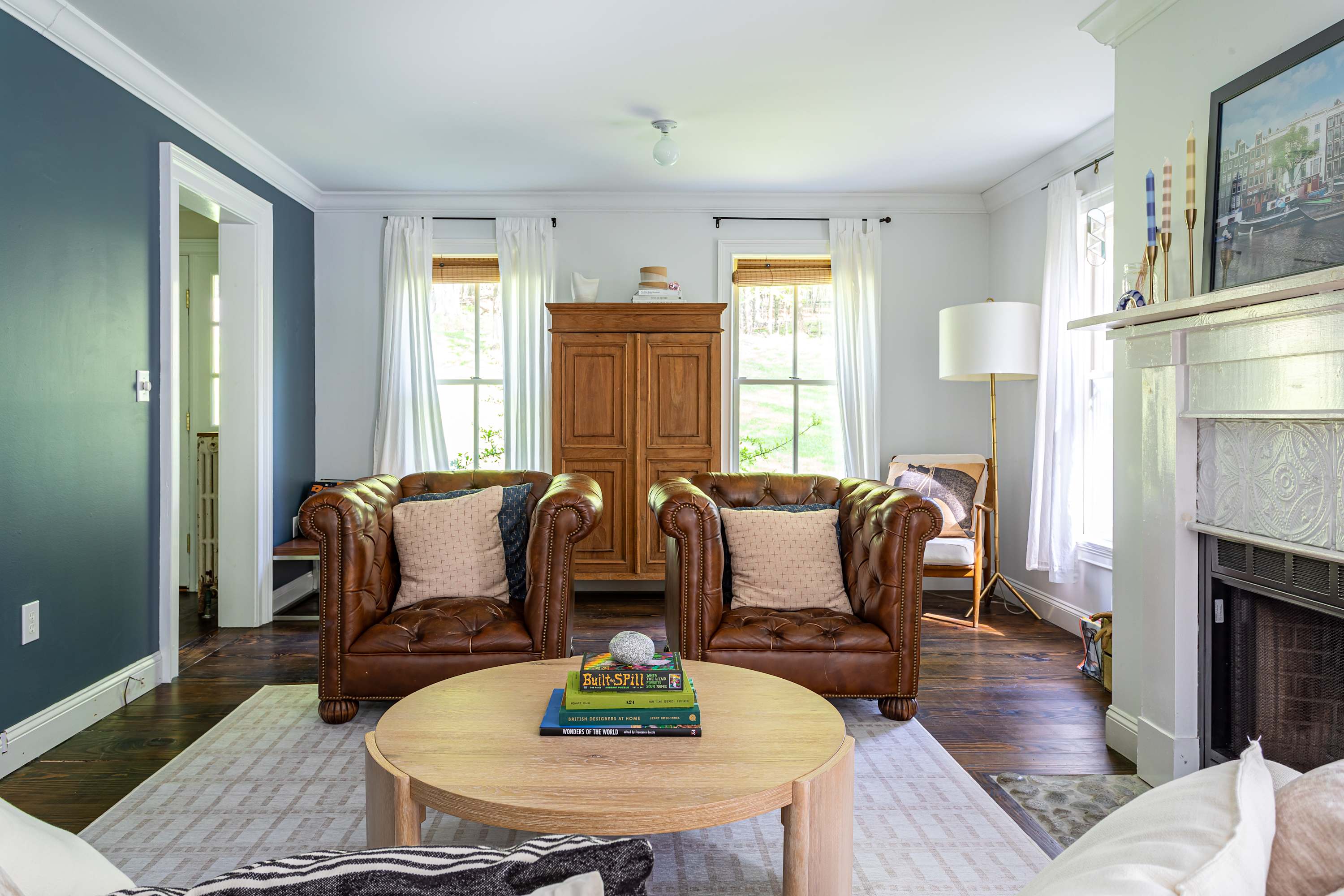 Elegant living room featuring two brown leather tufted armchairs, a white fireplace, and a round wooden coffee table. White walls and dark hardwood floors create a classic, inviting atmosphere.