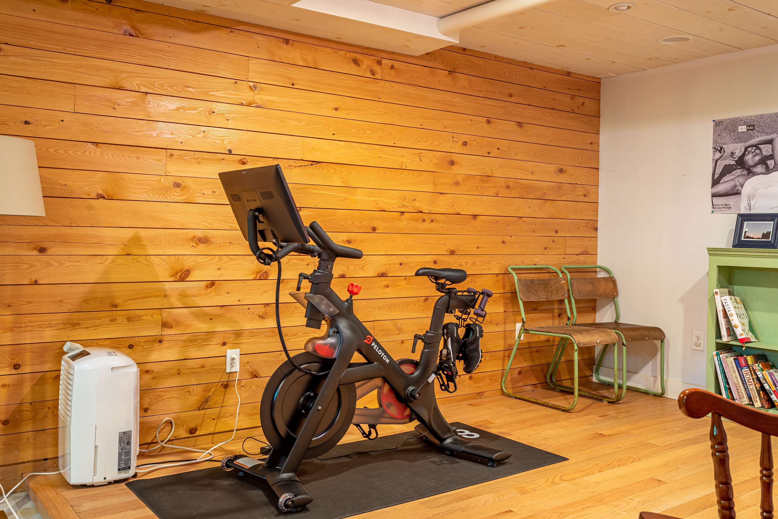 Dedicated fitness area within a wood-paneled recreation room, featuring a black Peloton exercise bike on a mat. A dehumidifier is visible in the corner, and a small green bookshelf with books adds a touch of color.