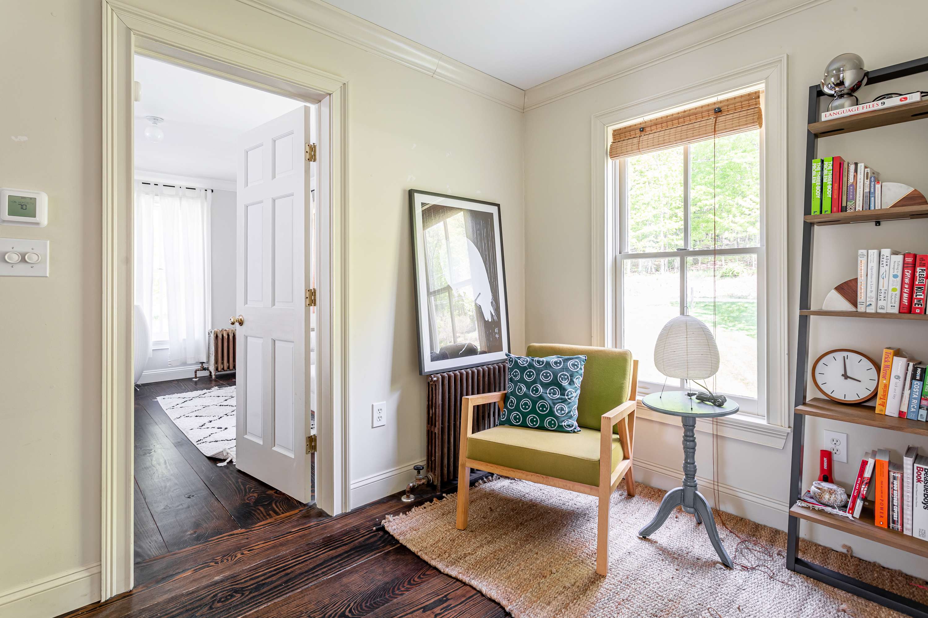 A cozy reading nook or office space with dark hardwood floors, featuring a green armchair, a small side table, and a tall bookshelf filled with books. A window with bamboo blinds provides natural light.
