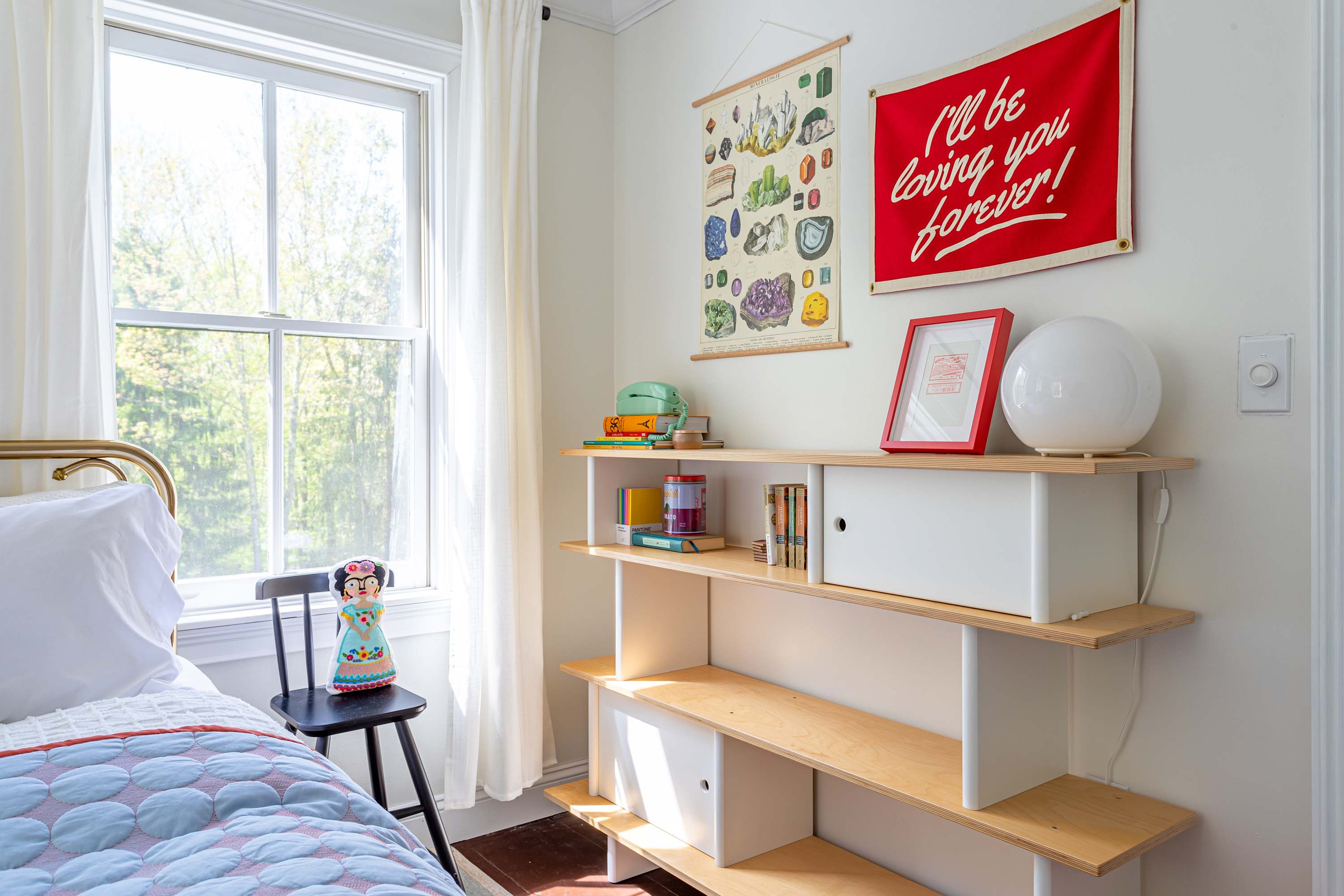 A close-up view of the bedroom's built-in shelving unit, adorned with decorative items and books, next to a window with white curtains. A red banner and a botanical chart hang above the shelves.