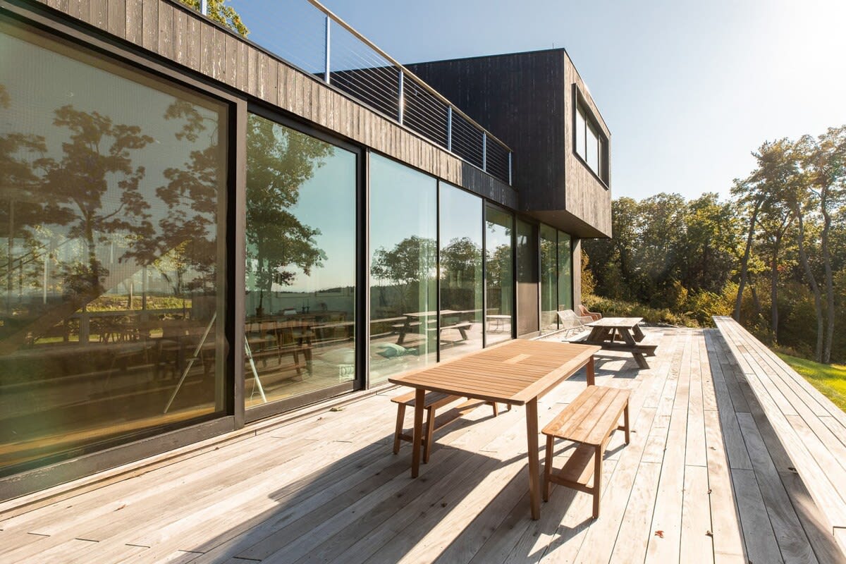 An expansive outdoor wooden deck with several picnic tables and benches, set against a modern dark-sided house with large glass sliding doors. Lush trees are visible in the background.