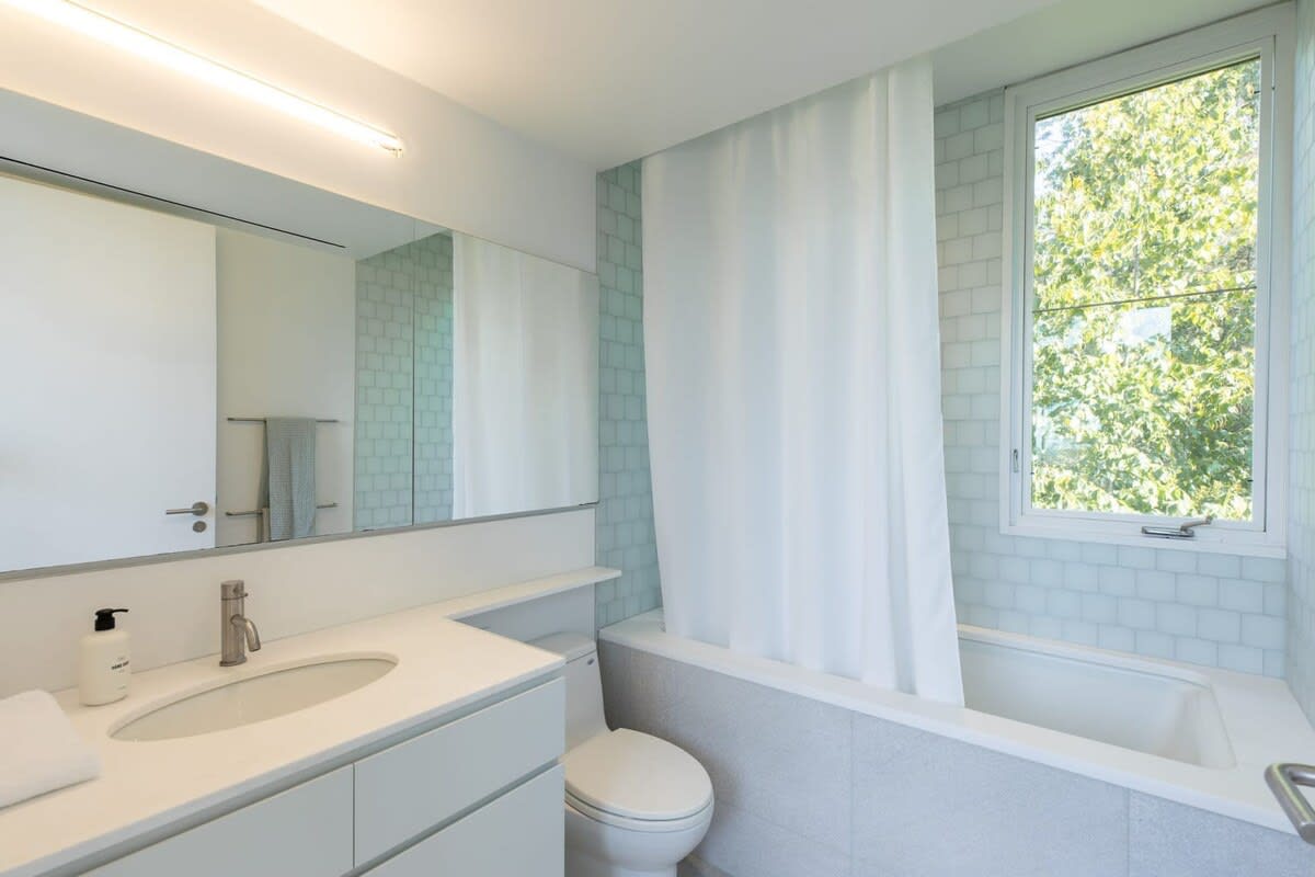 A modern bathroom with a white vanity, sink, and toilet, featuring light blue subway tiles around the bathtub. A large window provides natural light and a view of trees.