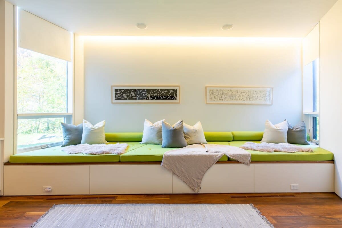 Inviting built-in bench seating area with vibrant green cushions and an array of throw pillows, featuring integrated storage cabinets below and subtle recessed lighting above, flanked by windows.