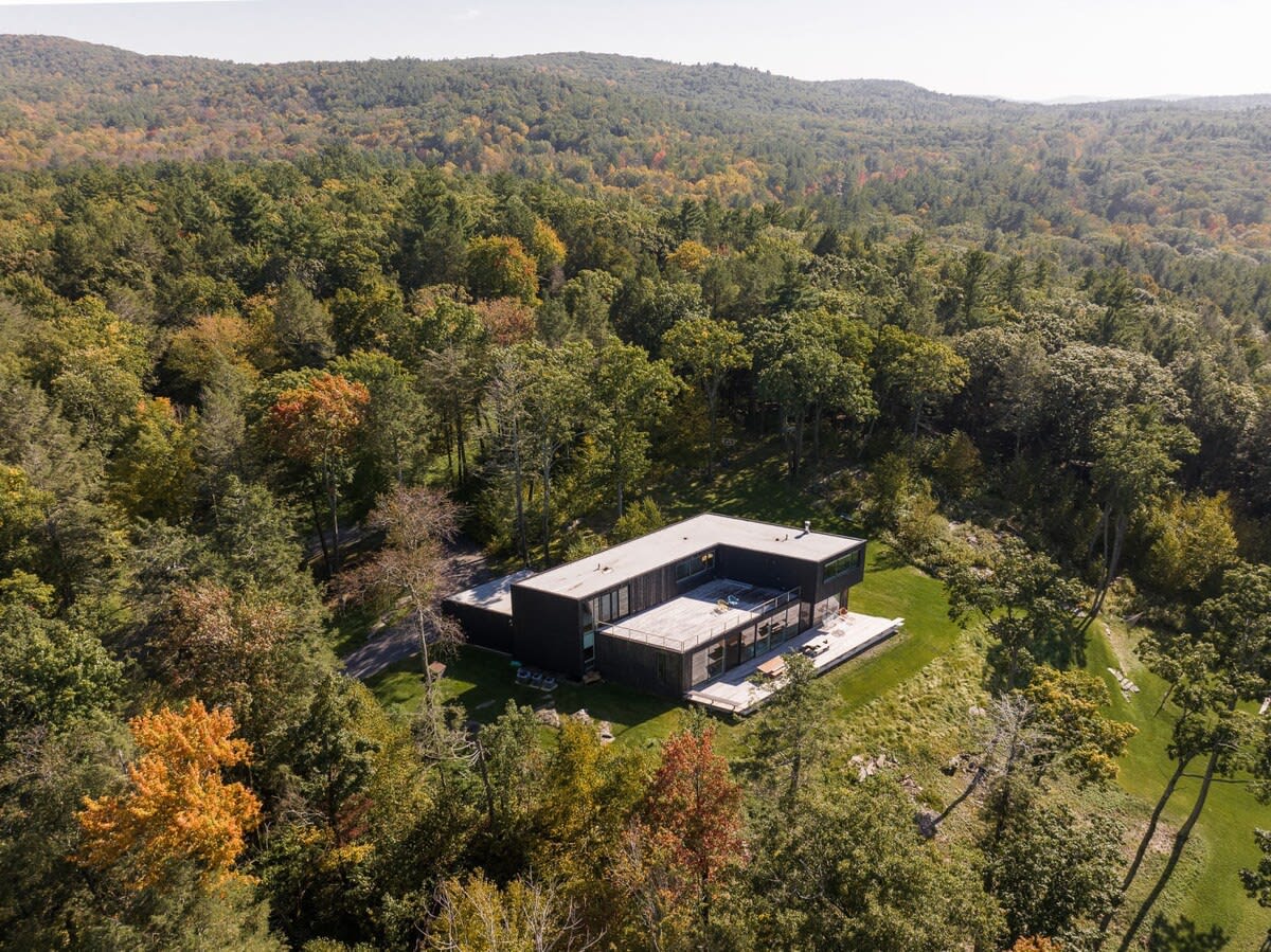 An aerial view showcasing a modern, dark-colored house with a flat roof, nestled within a vast, vibrant forest displaying autumn colors. The property includes a large lawn area.