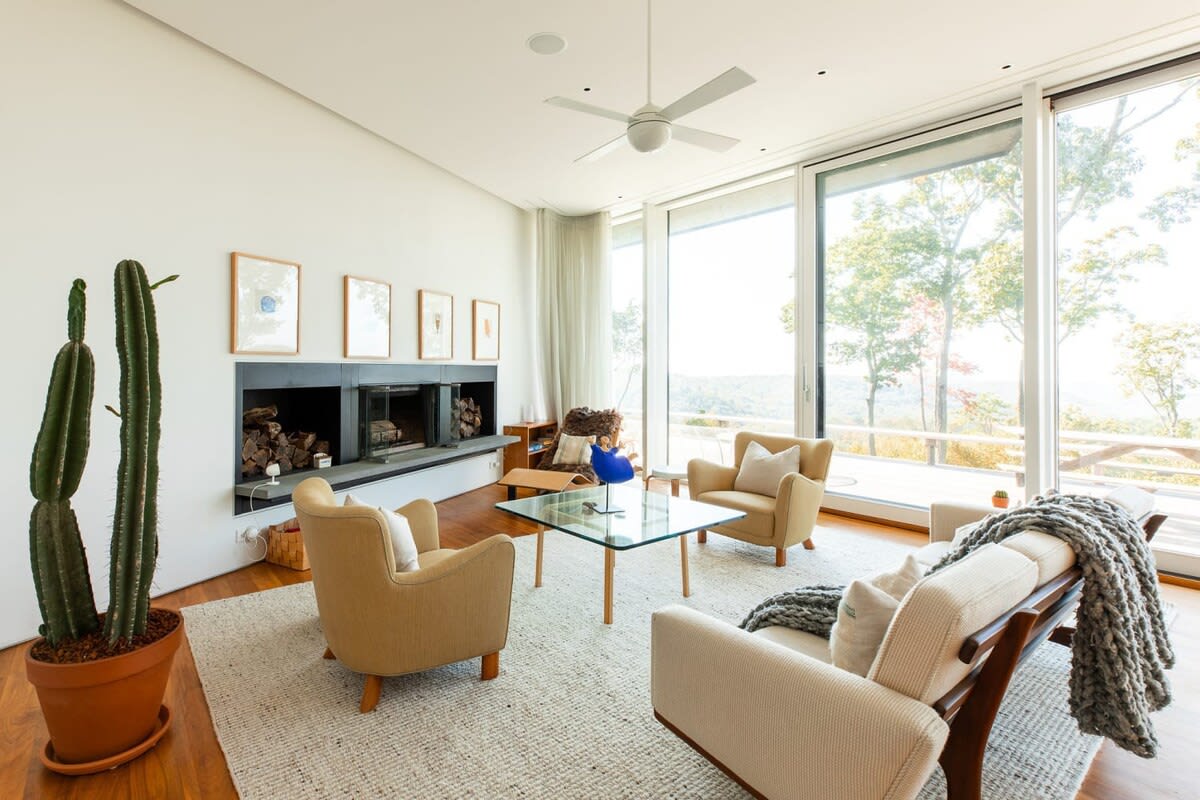 Spacious living room with a modern fireplace, a white sofa, two beige armchairs, a glass coffee table, and floor-to-ceiling windows overlooking a scenic landscape. A large cactus plant adds a touch of greenery.