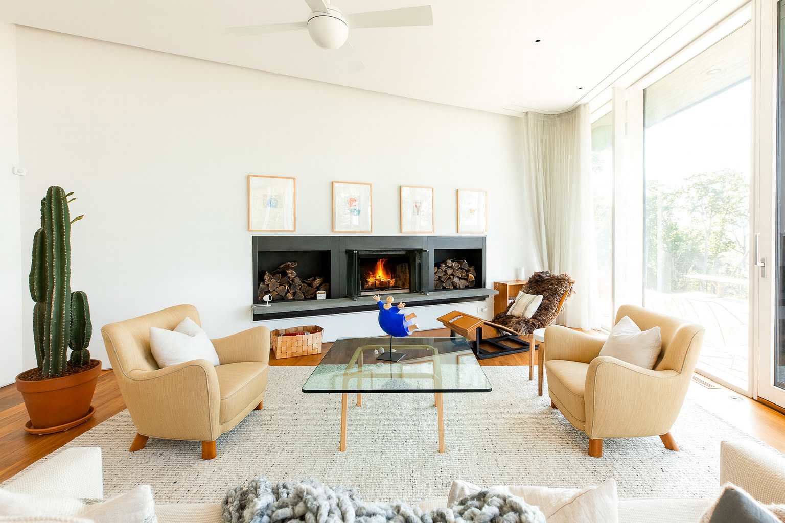 Bright living room with white walls, a modern fireplace with wood storage, two beige armchairs, a glass coffee table, and a tall cactus plant.