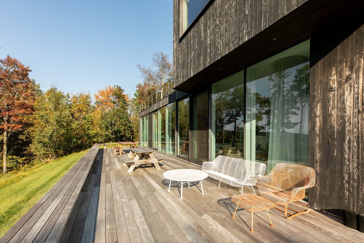 Wide wooden deck extending from a modern black house with floor-to-ceiling windows, featuring contemporary white and orange wireframe outdoor seating and a picnic table, blending indoor-outdoor living seamlessly.