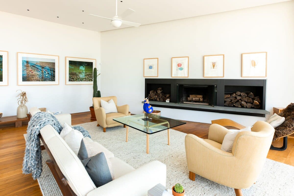 Cozy living room featuring a long white sofa, two beige armchairs, a glass coffee table, a modern fireplace with integrated wood storage, and framed artwork on the wall, creating a comfortable and stylish gathering space.