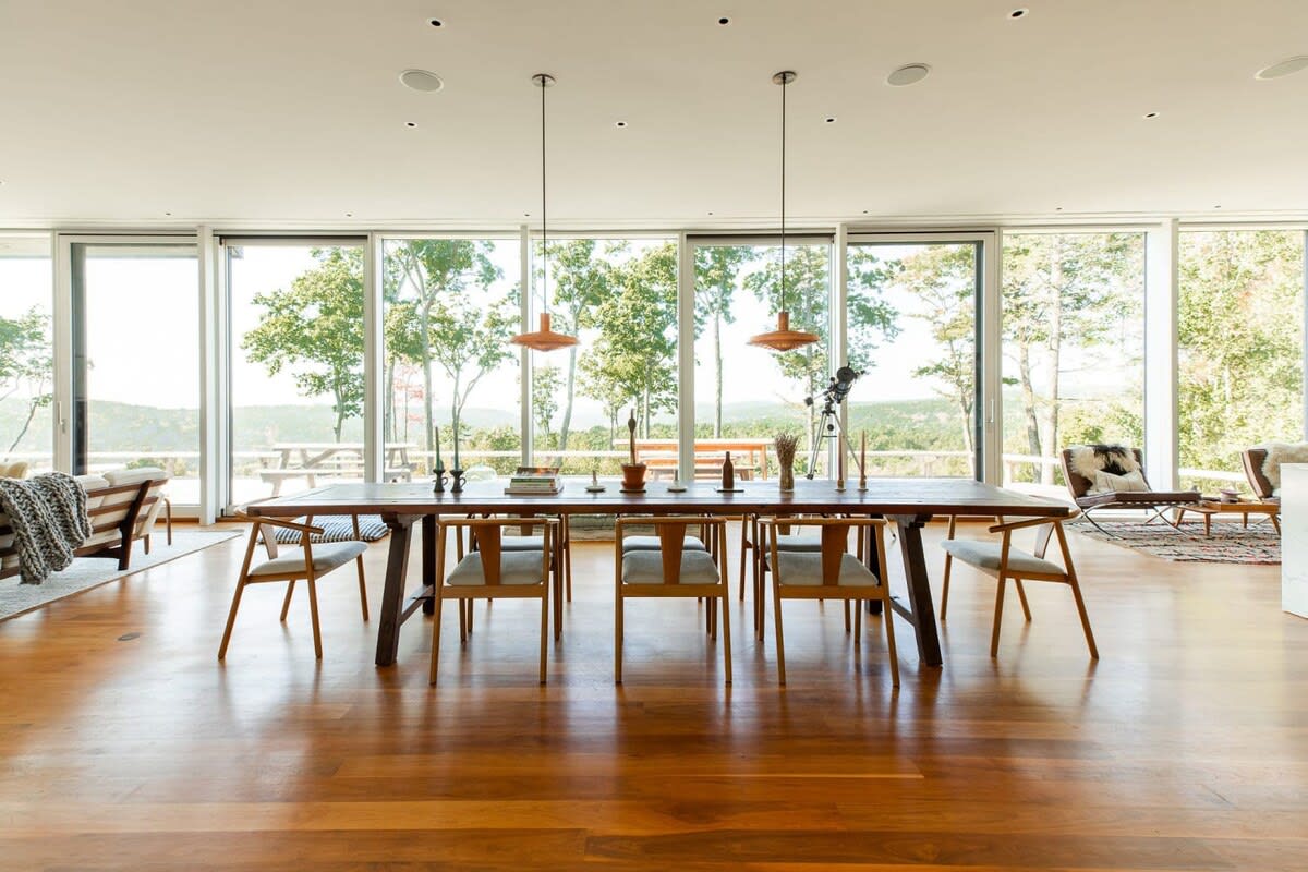 Bright dining area with a long wooden table and minimalist chairs, featuring floor-to-ceiling windows that provide abundant natural light and expansive views of the surrounding trees and landscape.