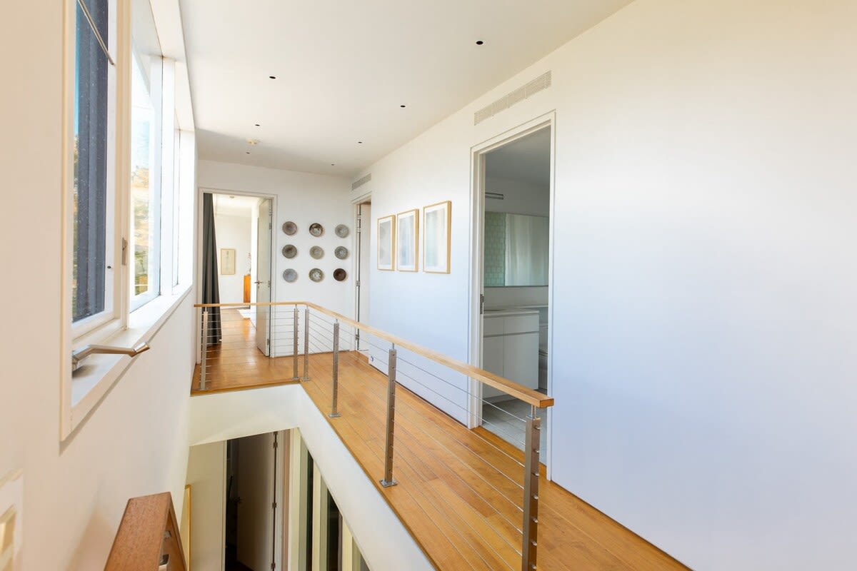 A bright, modern upper-level hallway with light hardwood floors and white walls, featuring a minimalist metal and wood railing overlooking a lower floor, with doorways leading to other rooms.