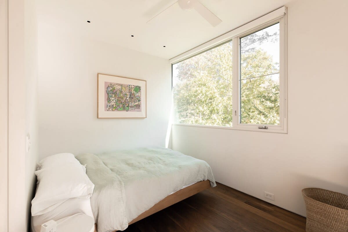 A minimalist bedroom featuring a low-profile bed with light green bedding, hardwood floors, and a large window offering views of lush trees. A small white nightstand is visible.