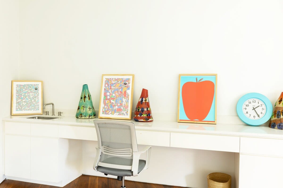 Dedicated office space with a long white built-in desk, an ergonomic office chair, and a small sink, decorated with vibrant framed artwork and a blue wall clock.
