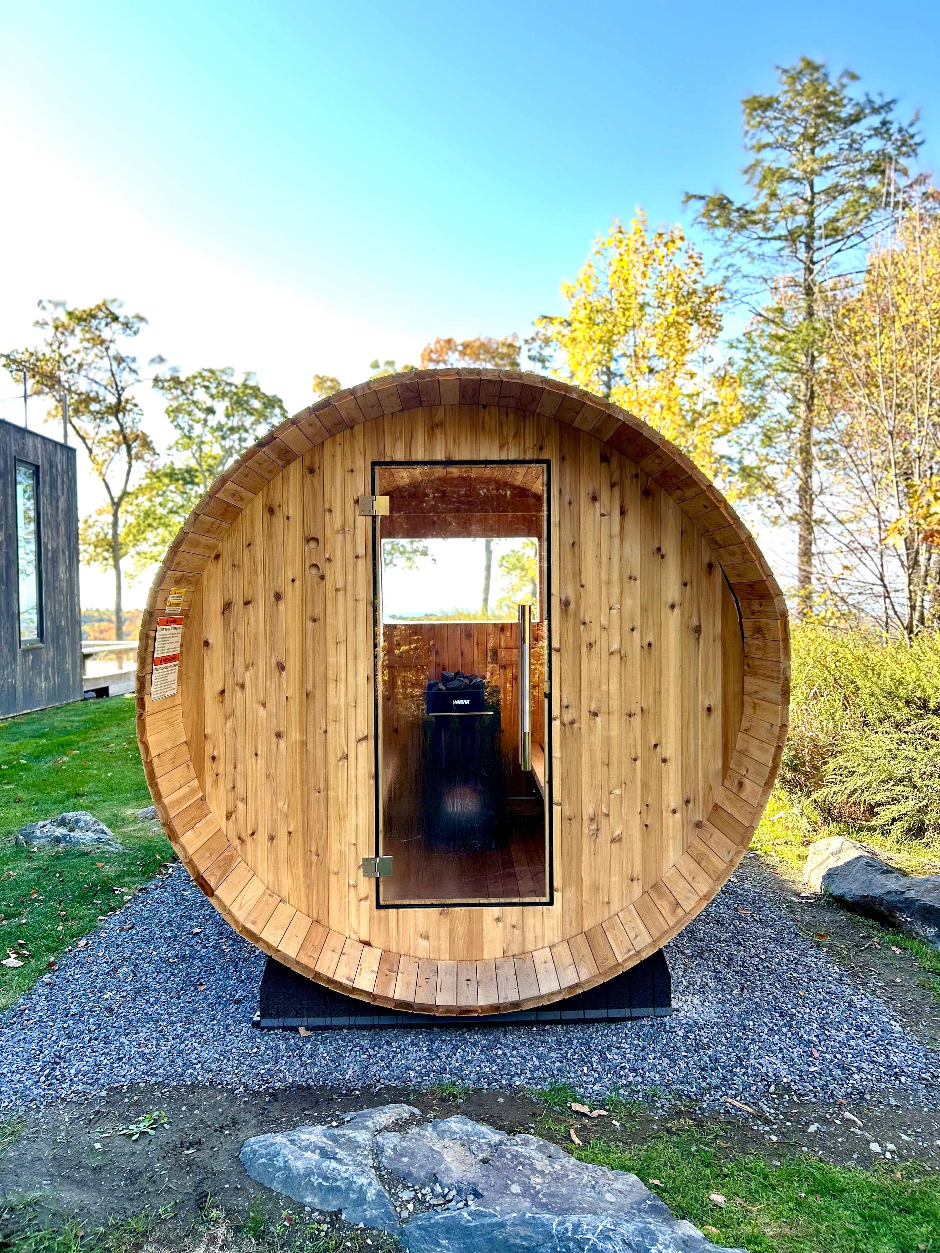 A unique barrel-shaped wooden sauna with a glass door, situated outdoors on a gravel patch. The surrounding landscape features green grass and autumn trees under a clear sky.