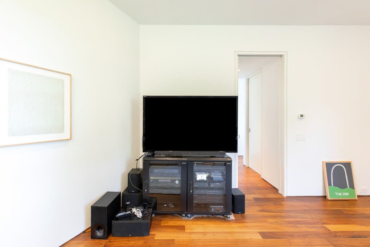 Entertainment corner featuring a large flat-screen TV on a media console with speakers, set against a white wall with minimalist artwork, on warm hardwood floors.