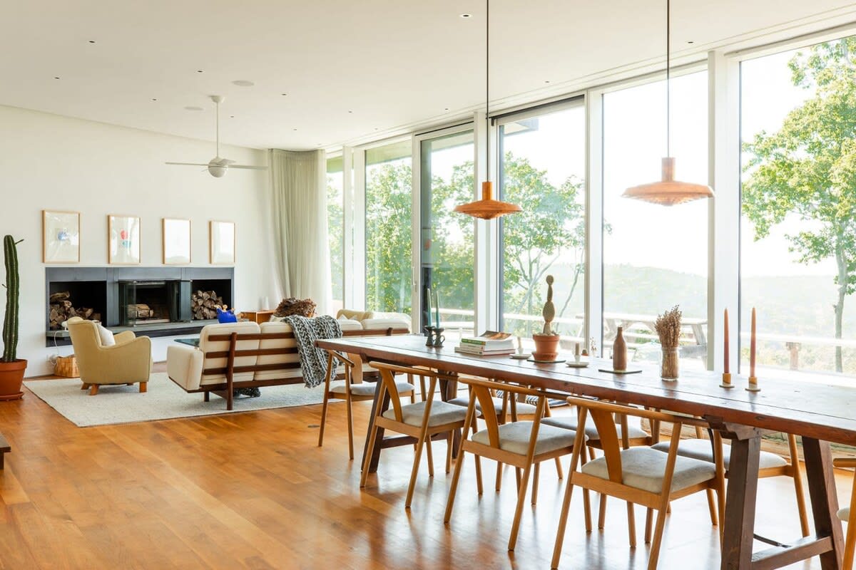 Spacious dining area featuring a long rustic wooden table with modern chairs, illuminated by two pendant lights, adjacent to a cozy living room with a fireplace and large windows overlooking nature.