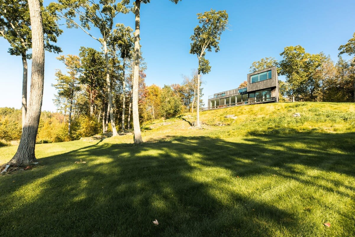 A modern, dark-clad house with large windows is nestled on a grassy hillside, surrounded by a mix of green and autumnal trees. The property features a spacious lawn and a clear blue sky overhead.