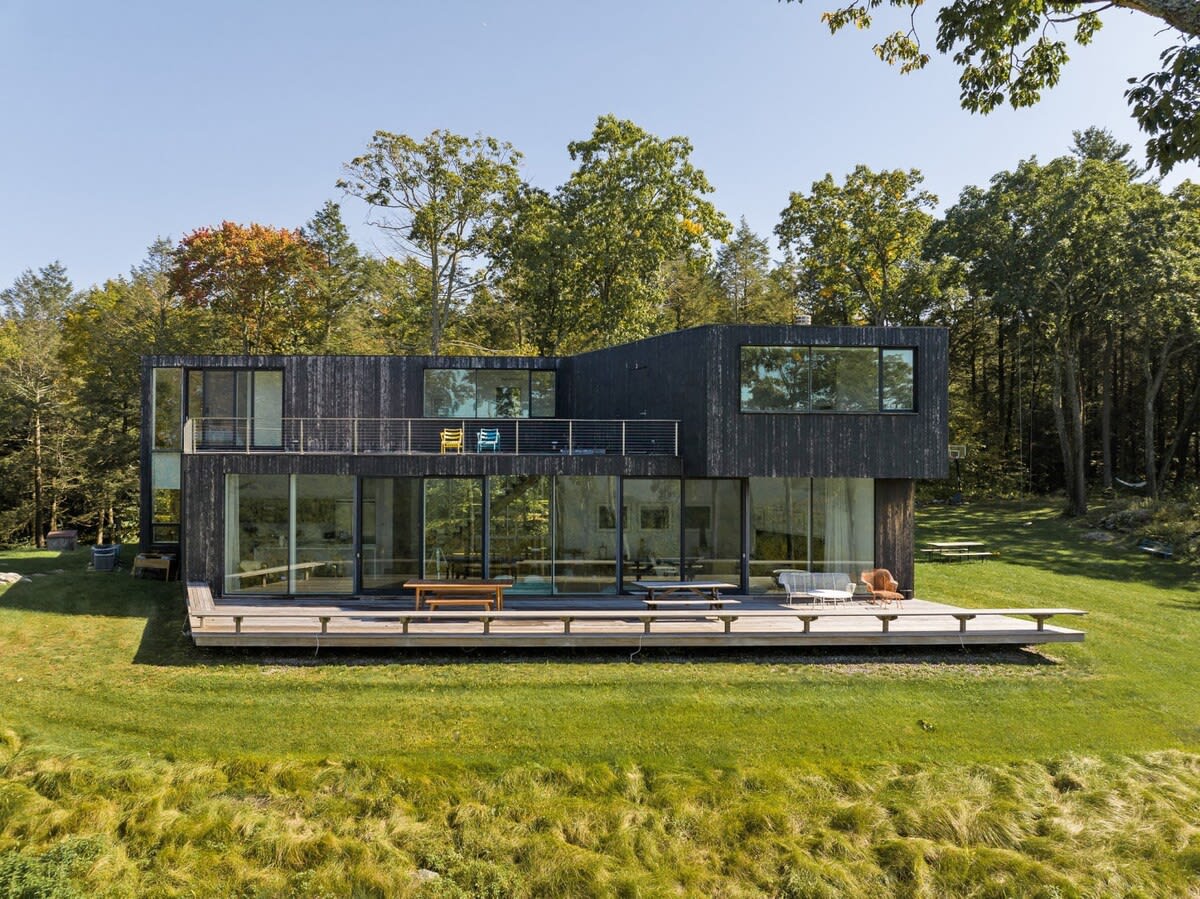 Modern black-clad house with large floor-to-ceiling windows and a wide wooden deck, surrounded by lush green grass and mature trees under a clear blue sky.