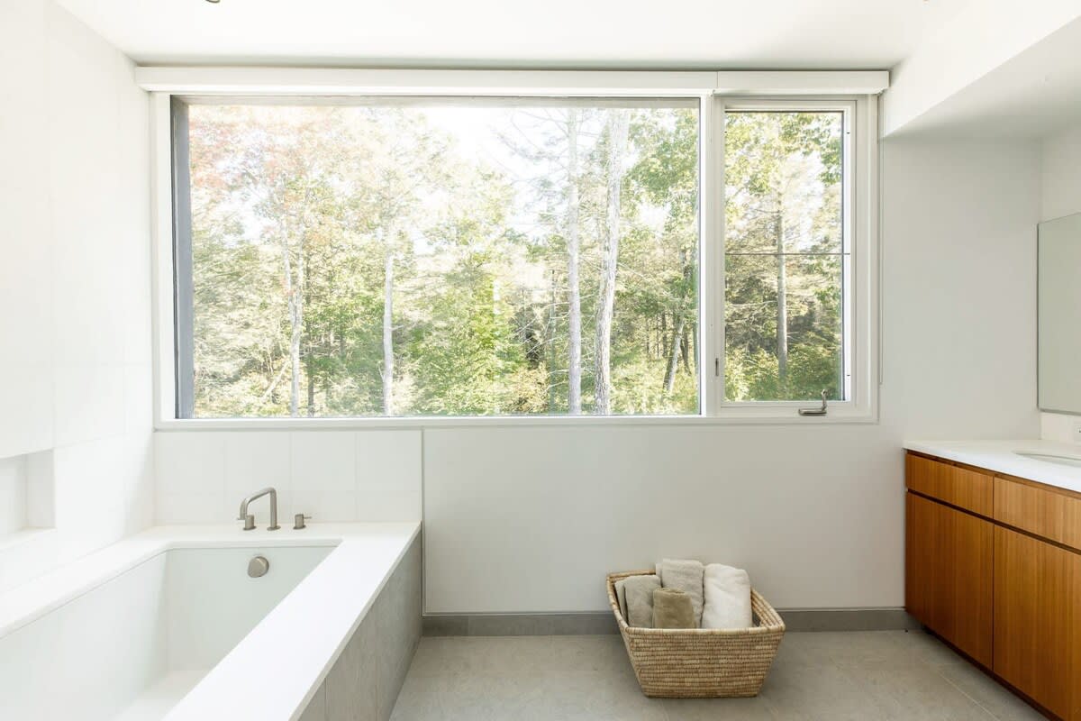 Modern bathroom with a built-in white bathtub, large window offering forest views, a wooden vanity with a sink, and a woven basket with towels, providing a bright and natural feel.
