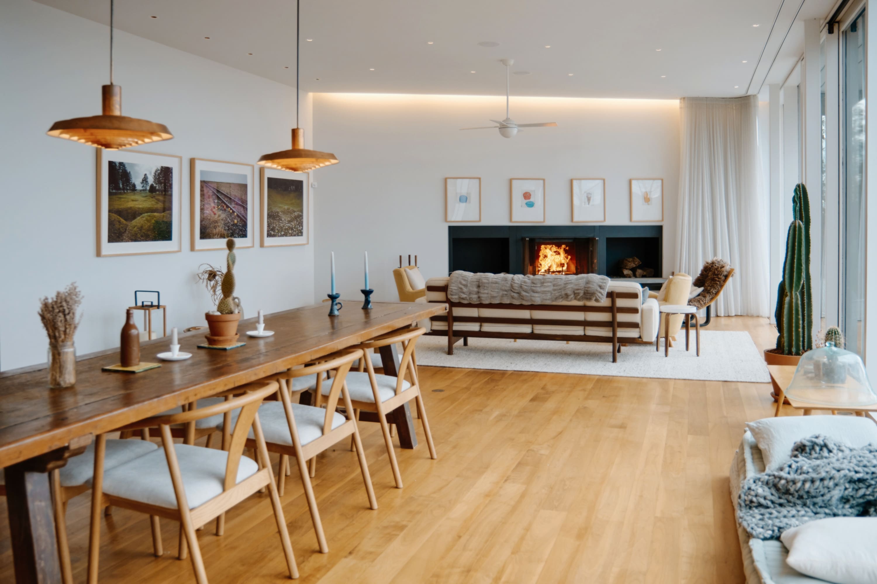 Open-concept living and dining area with a long wooden dining table and chairs, leading to a bright living space with a fireplace, sofa, and large windows.