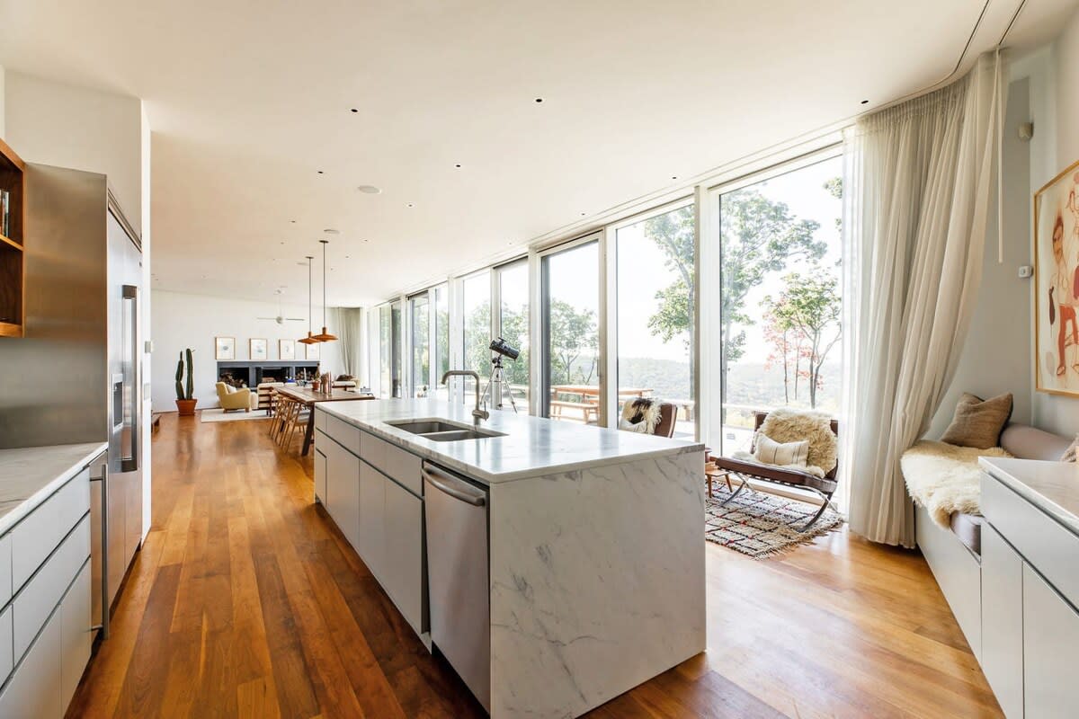 Expansive open-concept living space with a large white marble kitchen island, leading to a dining area and living room, all framed by floor-to-ceiling windows offering panoramic outdoor views.