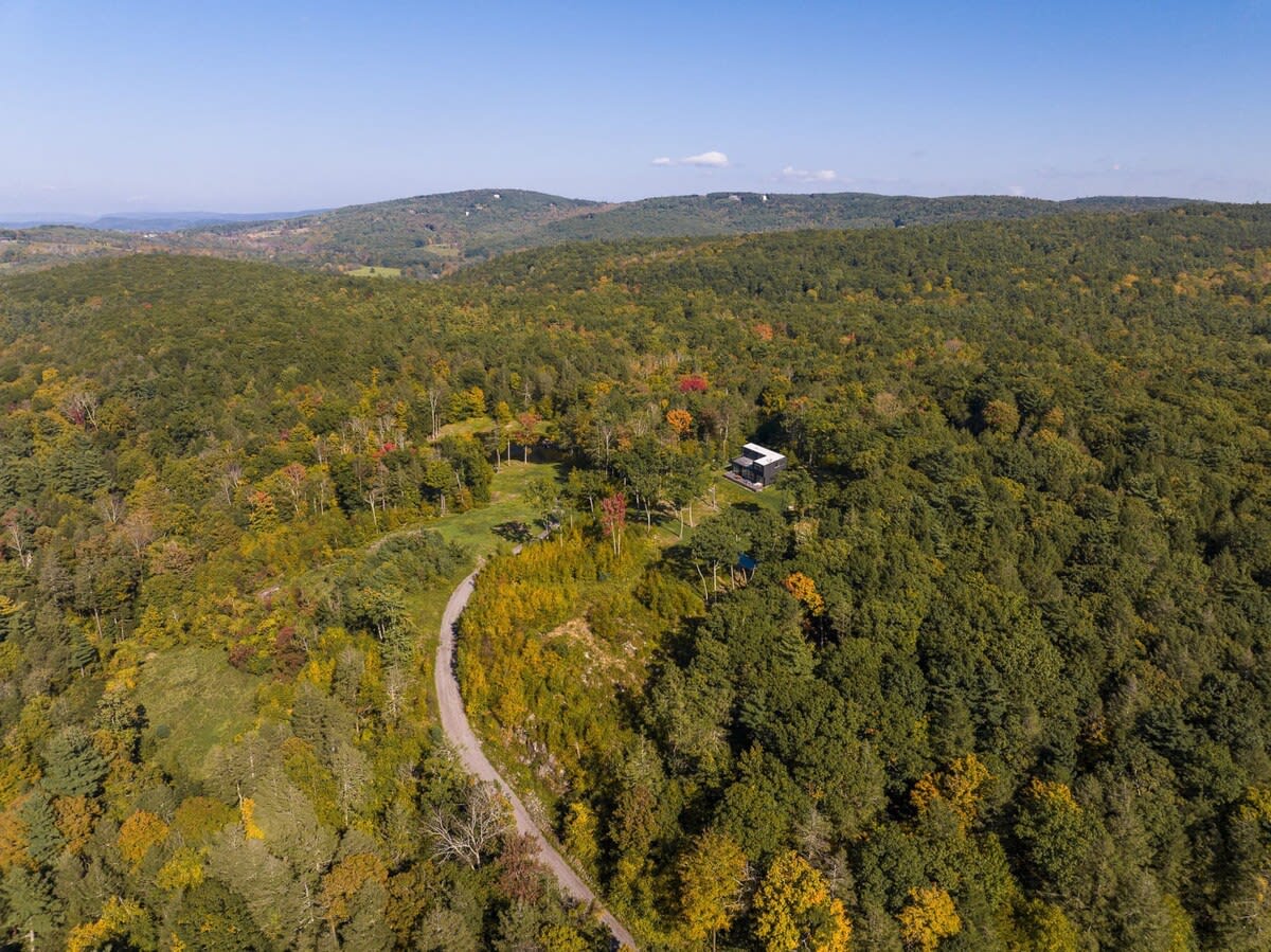 A sweeping aerial view captures the modern house nestled within a vast, vibrant autumn forest, showcasing a winding road and distant rolling hills. The landscape is ablaze with fall colors under a clear blue sky.