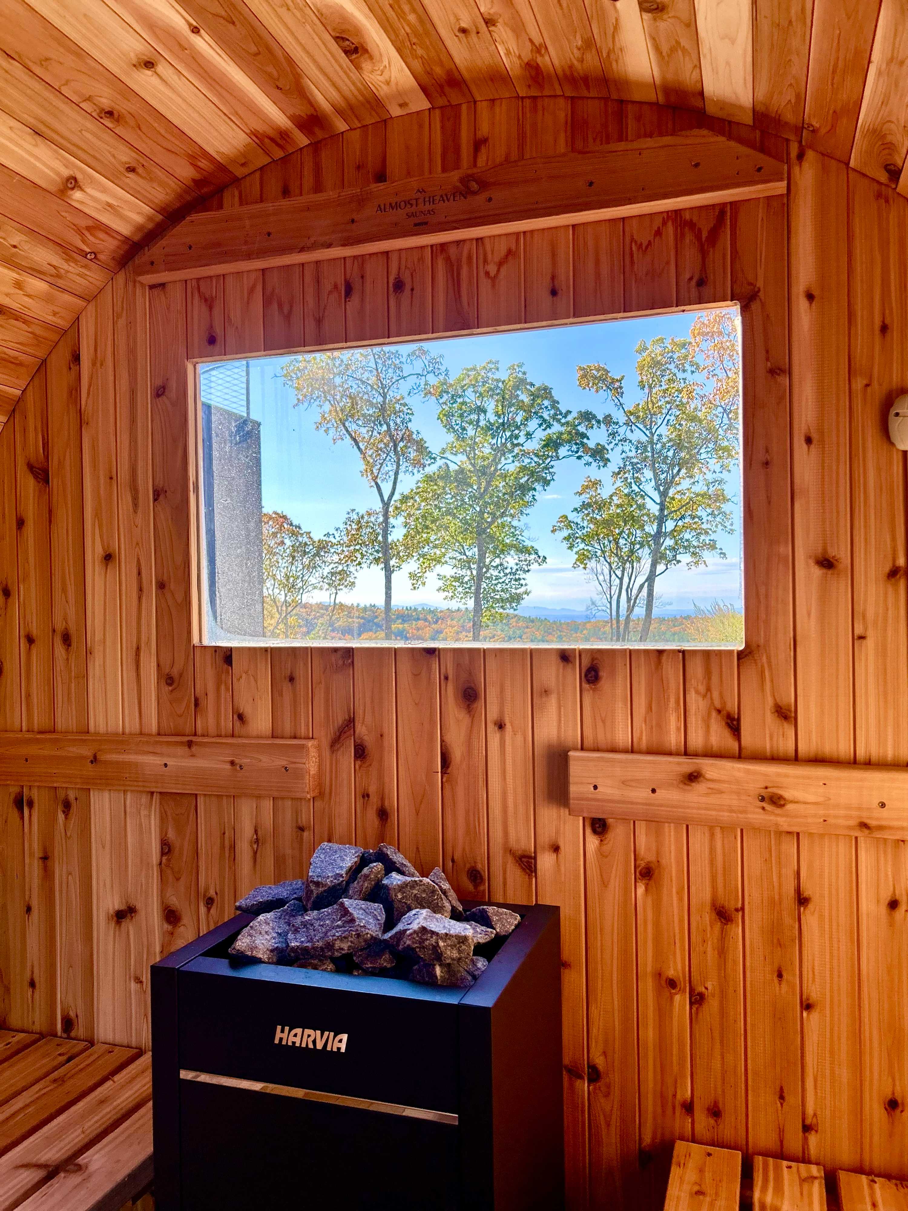 Interior of a wooden barrel sauna with a window offering views of trees and the distant landscape, featuring a modern electric sauna heater with rocks.