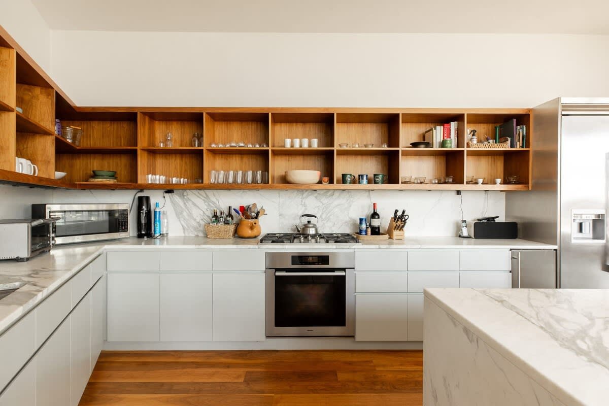 Detailed view of the modern kitchen with light wood open shelving, white marble countertops and backsplash, a stainless steel gas range and oven, and light gray lower cabinets, all on warm hardwood floors.