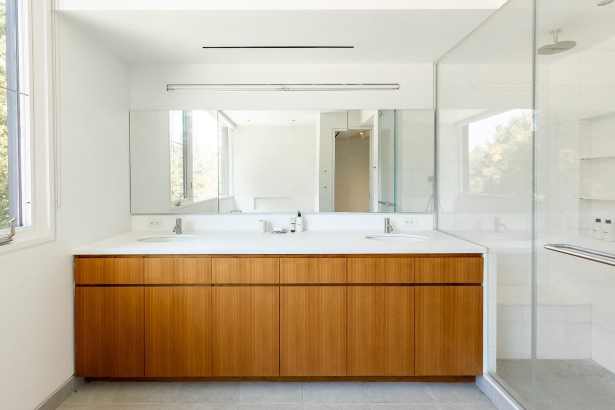 A bright, contemporary bathroom featuring a long wooden double vanity with white countertops and two sinks, a large mirror, and a glass-enclosed shower visible to the right.