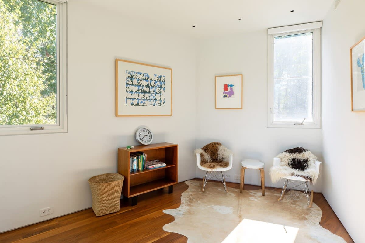 A bright room with hardwood floors, two large windows, and white walls adorned with colorful abstract art. It features a low wooden bookshelf, two modern chairs with furry throws, and a cowhide rug.