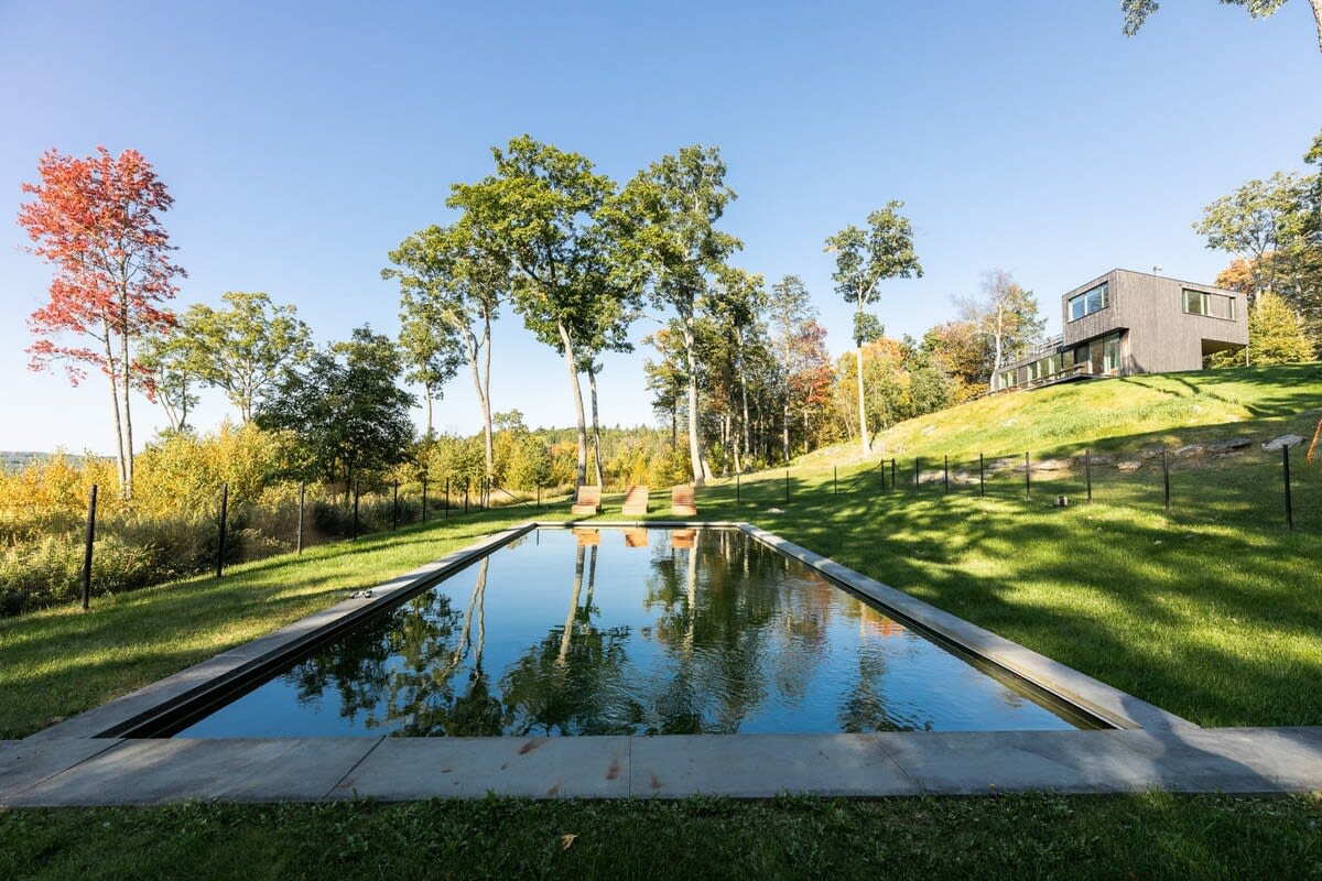 Rectangular swimming pool with dark water reflecting the sky and surrounding trees, set in a grassy lawn with a modern house visible on a hill in the background.
