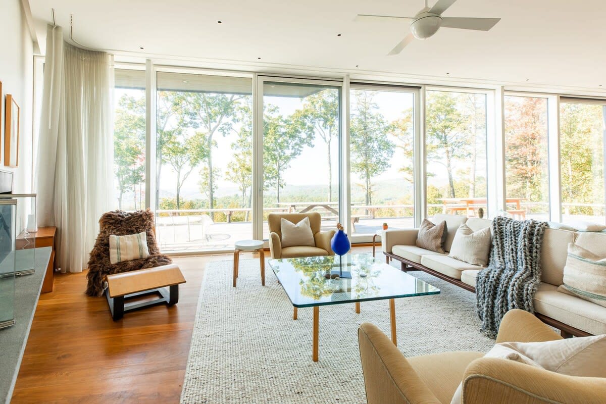Spacious living room with floor-to-ceiling windows offering panoramic views of the surrounding landscape, furnished with a sofa, armchairs, and a glass coffee table.
