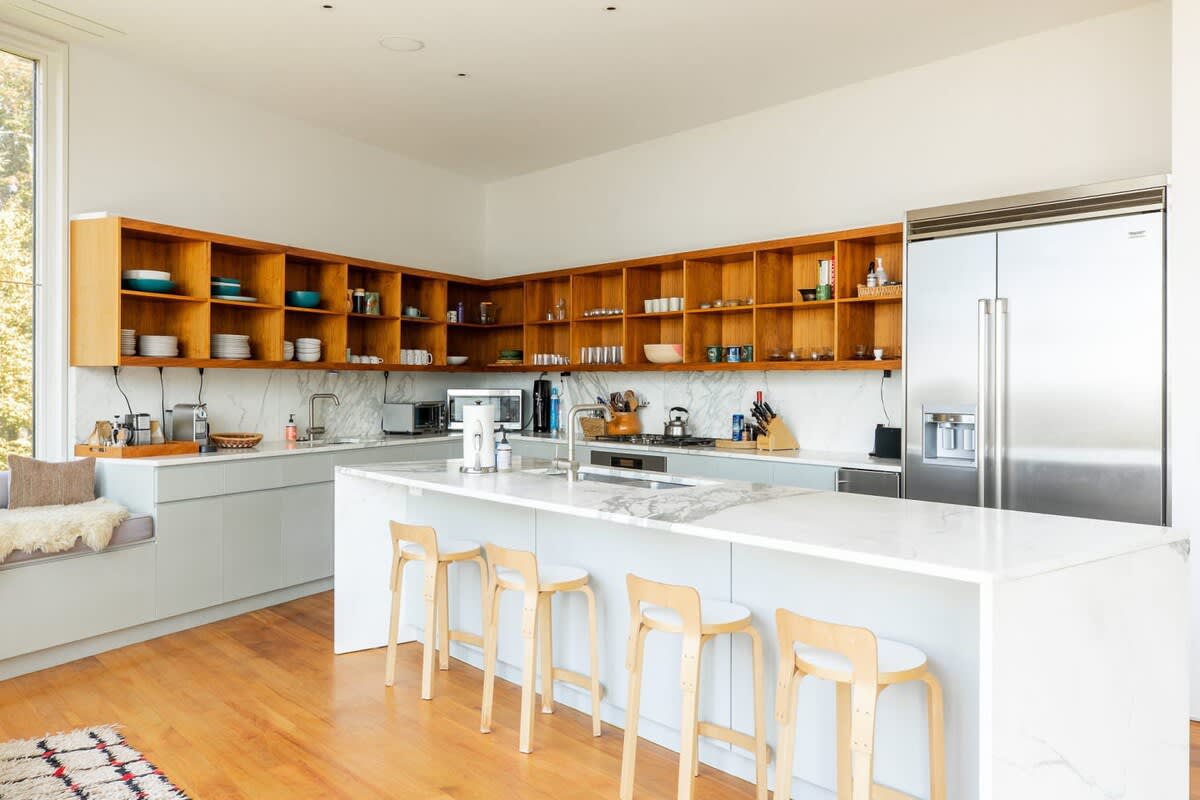 Bright, modern kitchen featuring light wood open shelving, white marble countertops, a large island with four light wood bar stools, and stainless steel appliances, all set on warm hardwood floors.