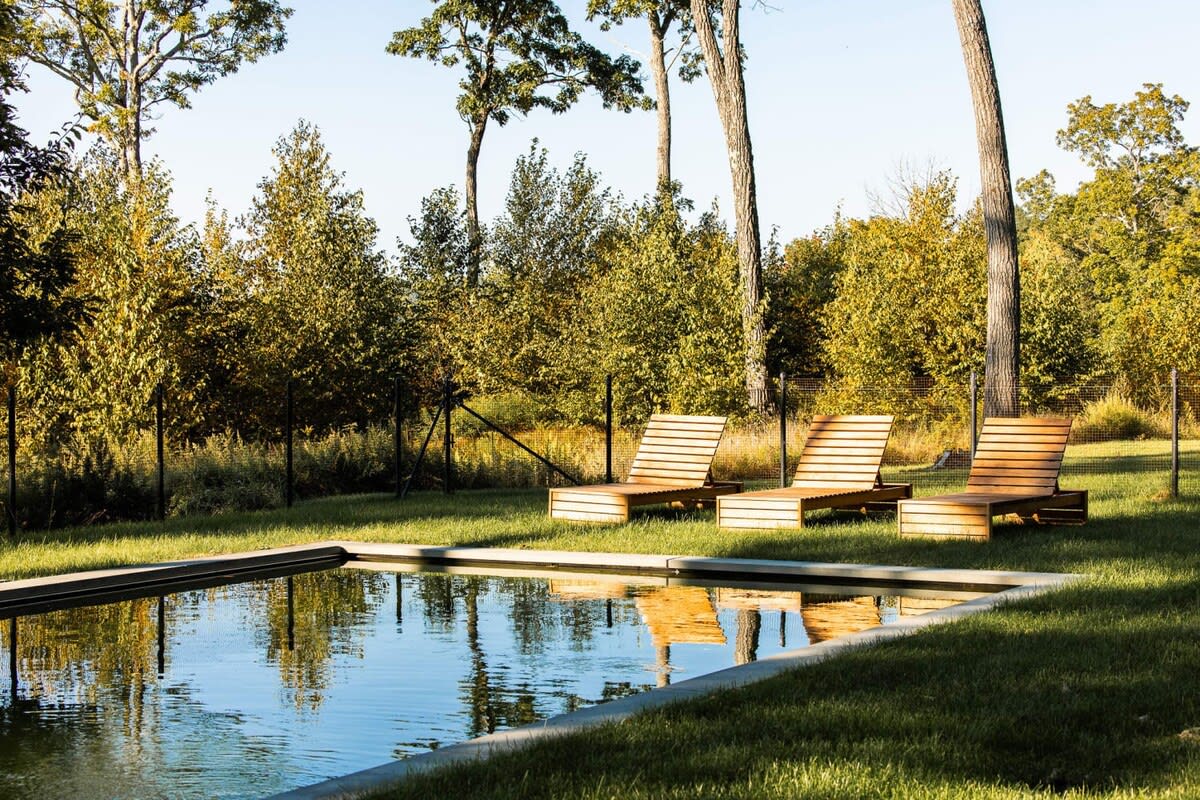 Rectangular swimming pool with three wooden lounge chairs on the grassy edge, surrounded by trees with autumn foliage under a bright sky.