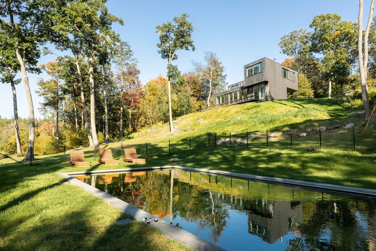 A rectangular swimming pool set in a grassy lawn, with wooden lounge chairs beside it. The modern dark-sided house is visible on a hill in the background, surrounded by trees.