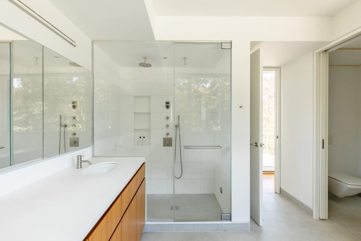 A modern bathroom with light gray tiled floors, a large glass-enclosed walk-in shower with multiple showerheads, and a wooden vanity with a white countertop and sink.