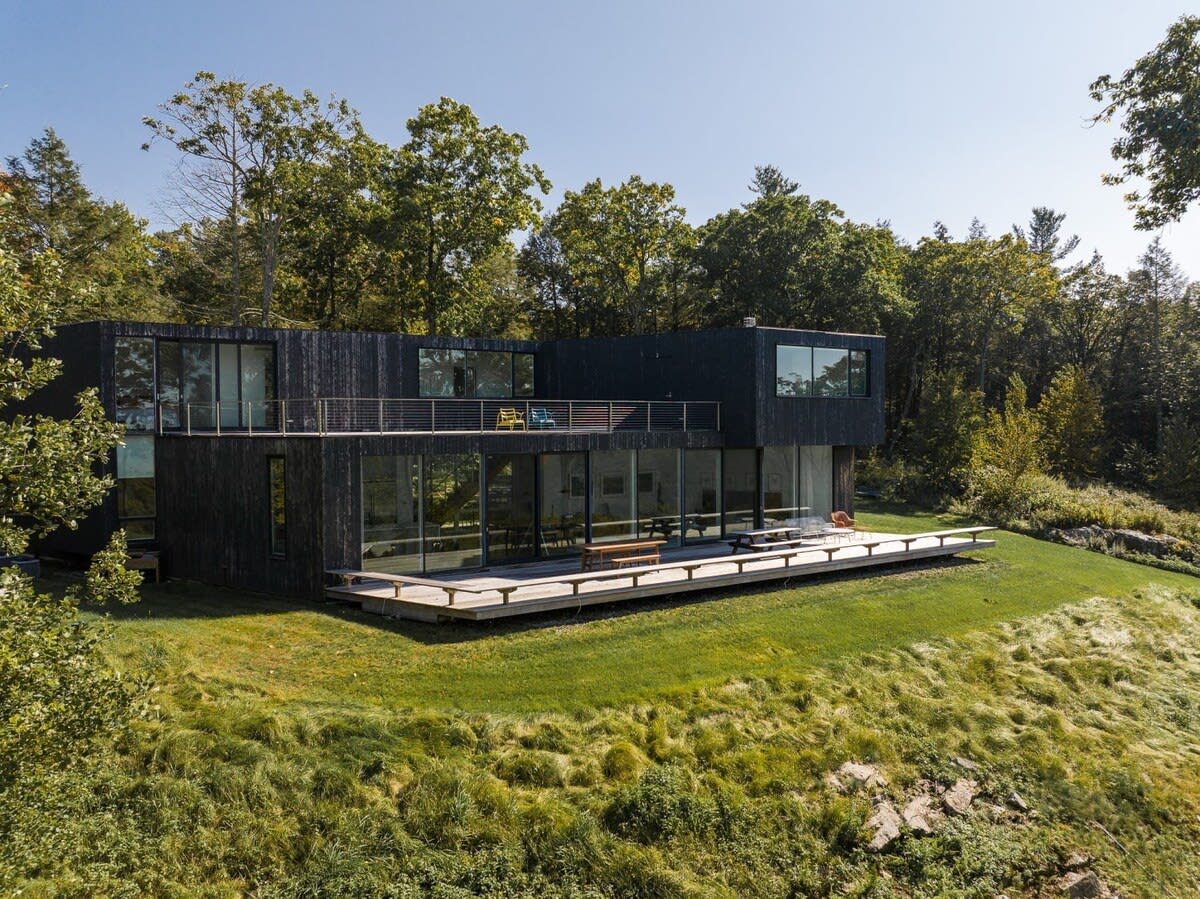 The striking modern house features a dark exterior with extensive floor-to-ceiling windows on both levels, opening onto a wide wooden deck. A second-story balcony with a metal railing offers additional outdoor space, all set against a lush green hillside.