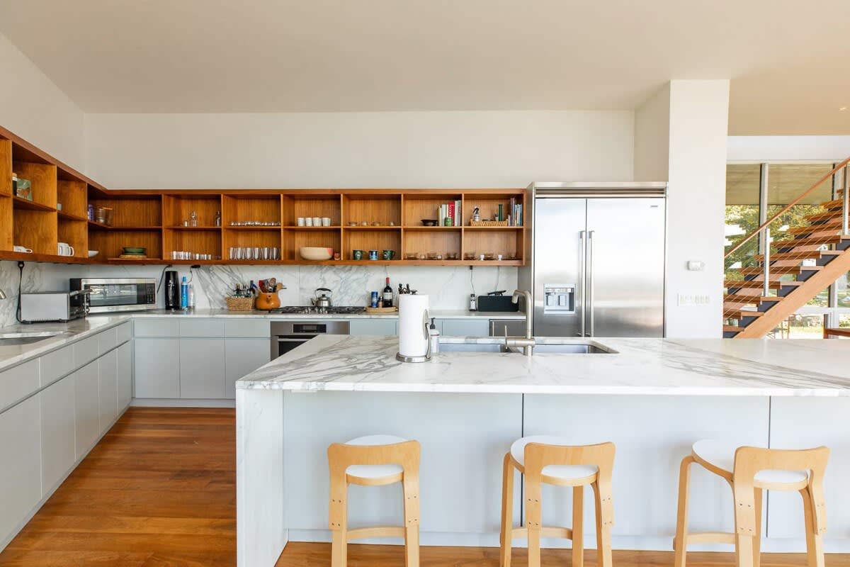 Bright, modern kitchen featuring light grey cabinetry, white marble countertops, a large island with bar stools, wooden open shelving, and stainless steel appliances. A wooden staircase is visible in the background, connecting to other living areas.