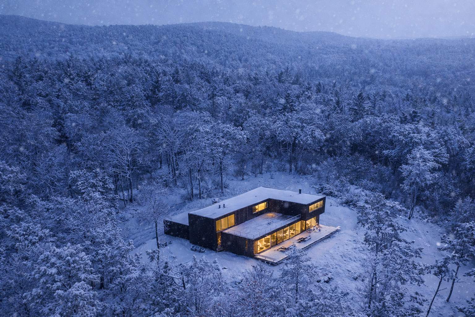 An aerial perspective reveals the modern house, with its warm interior lights, situated amidst a snow-covered forest under a soft, diffused winter sky. The surrounding trees are dusted with snow, highlighting the secluded, serene setting.